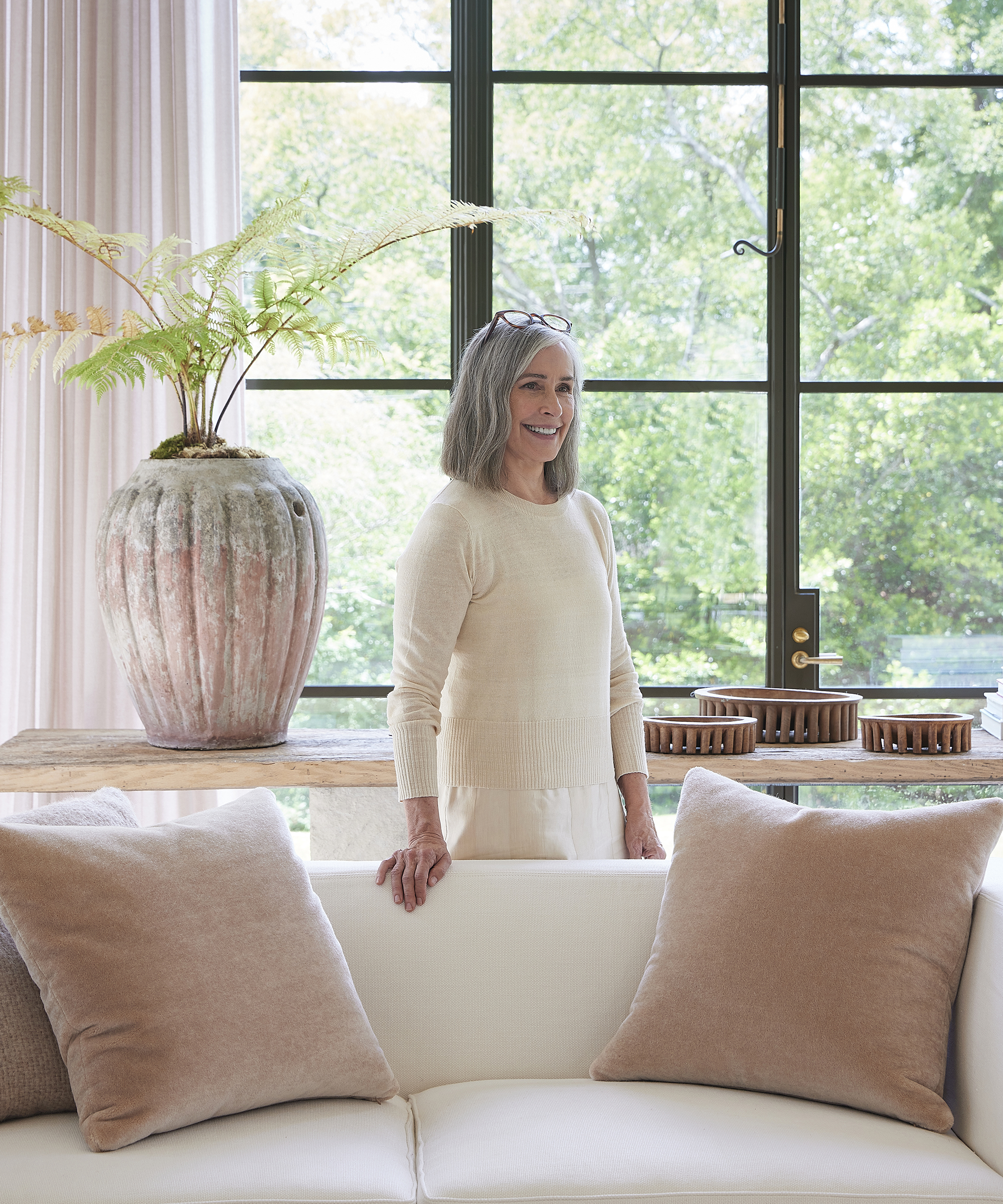 betsy brown stanging in a cream sweater in front of a white couch with taupe accent pillows in front of a large window