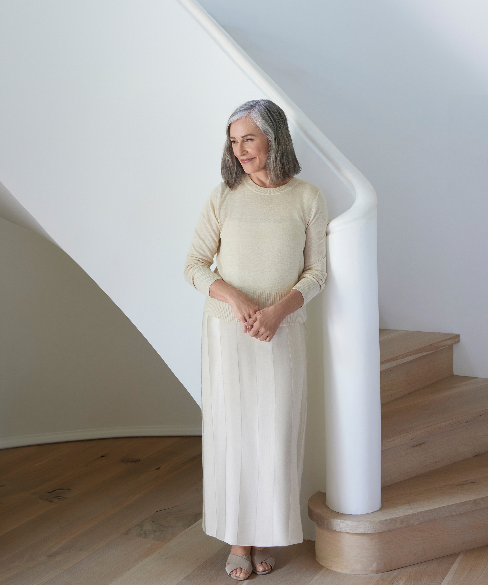 betsy brown standing in a creme pullover sweater and creme maxi dress in a white with oak floor stairwell