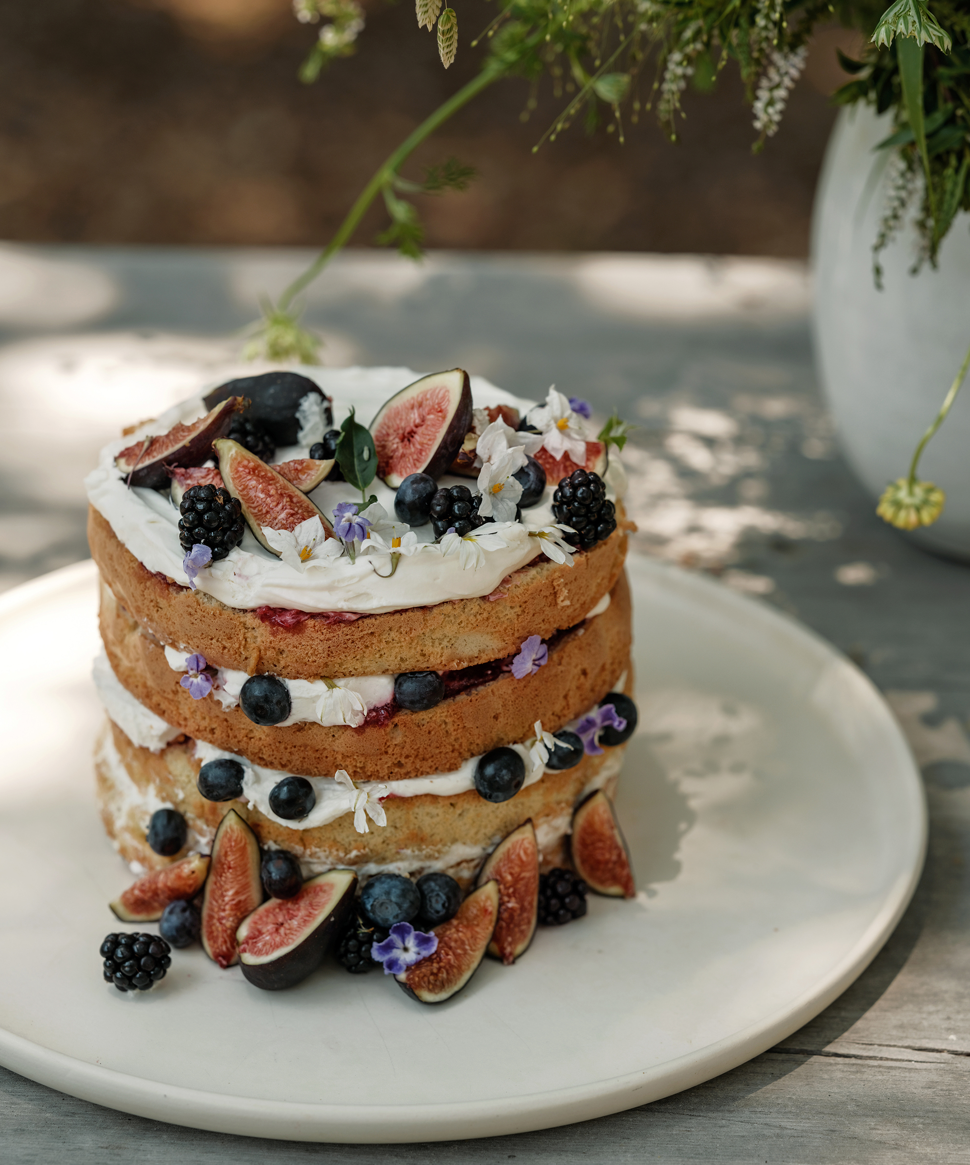 a layered berry cake on an ivory serving platter on an outdoor wood dining table