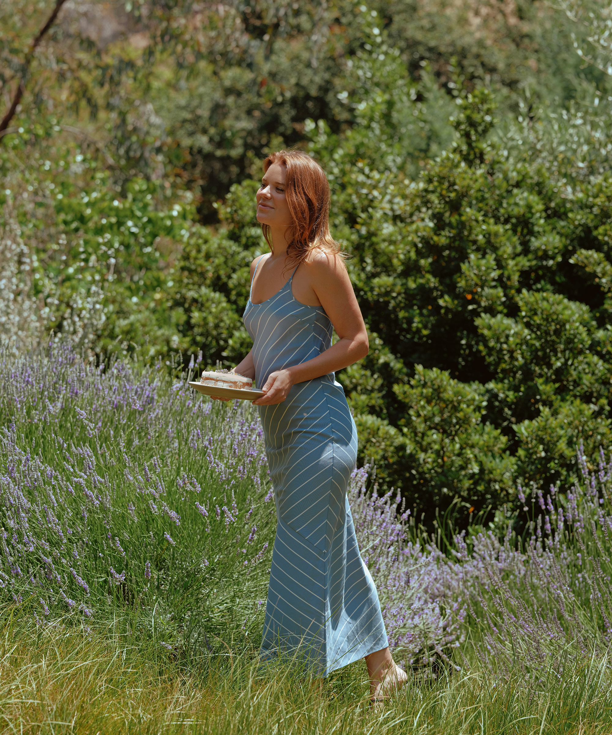 A woman in a long, blue, striped dress stands outdoors among tall grass and lavender, holding a plate of food inspired by vibrant recipes, with lush green foliage in the background.