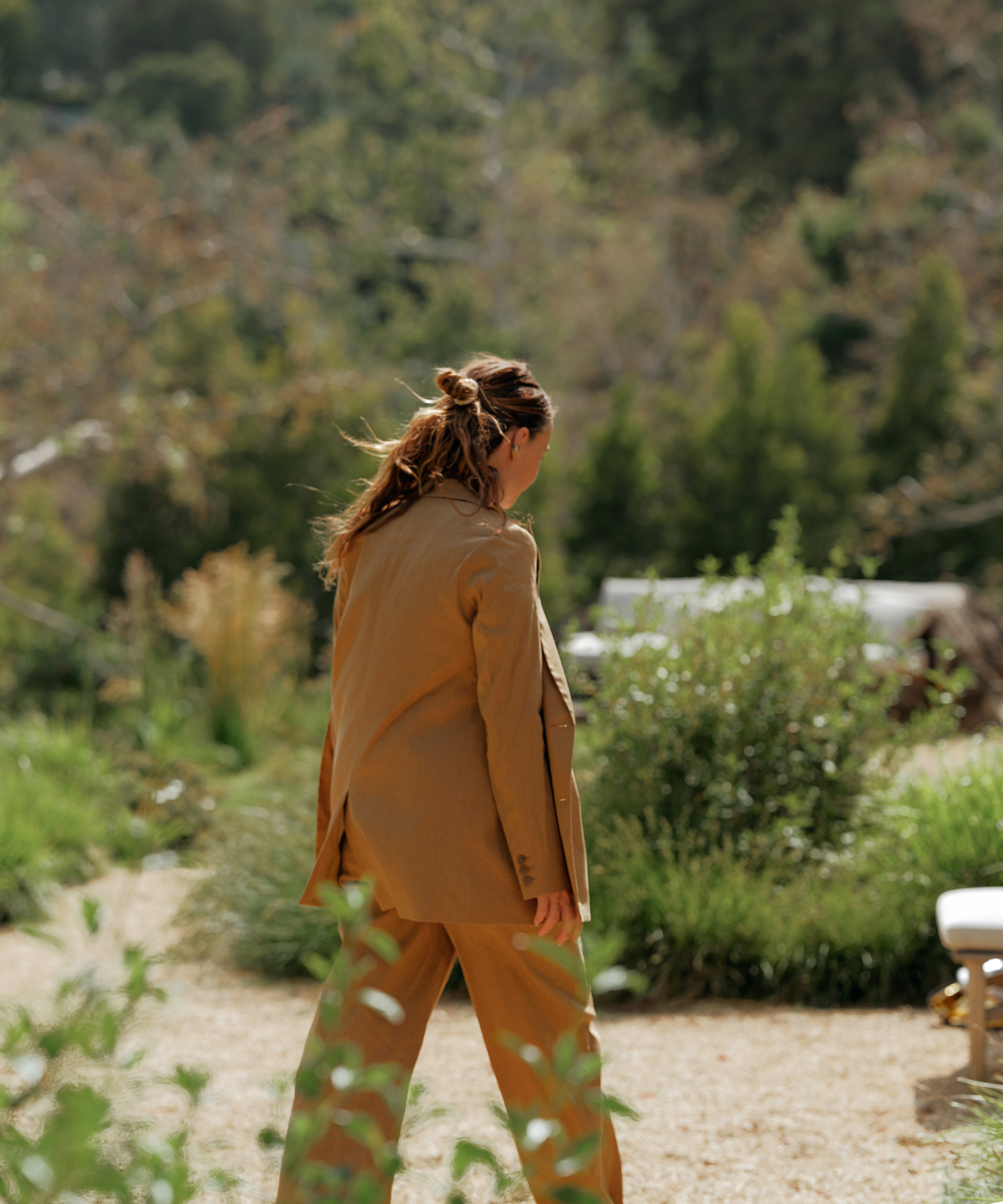 A person with long hair in a ponytail, wearing a tan pant suit, walks along a gravel path in a lush, green outdoor garden setting. Their back is to the camera.
