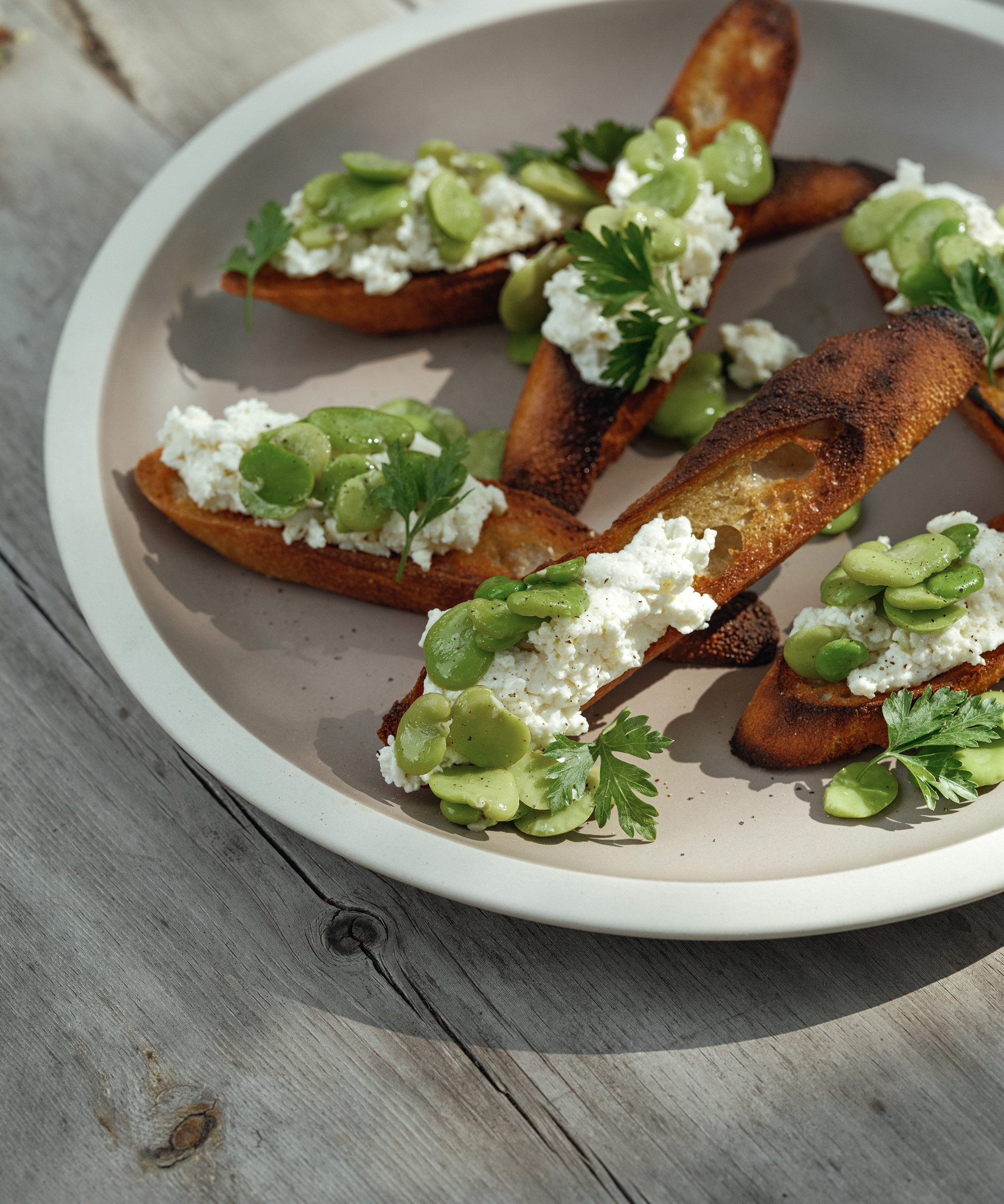 A plate of toasted baguette slices topped with ricotta cheese, broad beans, and fresh parsley, inspired by the flavors of La Morra, arranged on a light-colored plate set on a rustic wooden table.