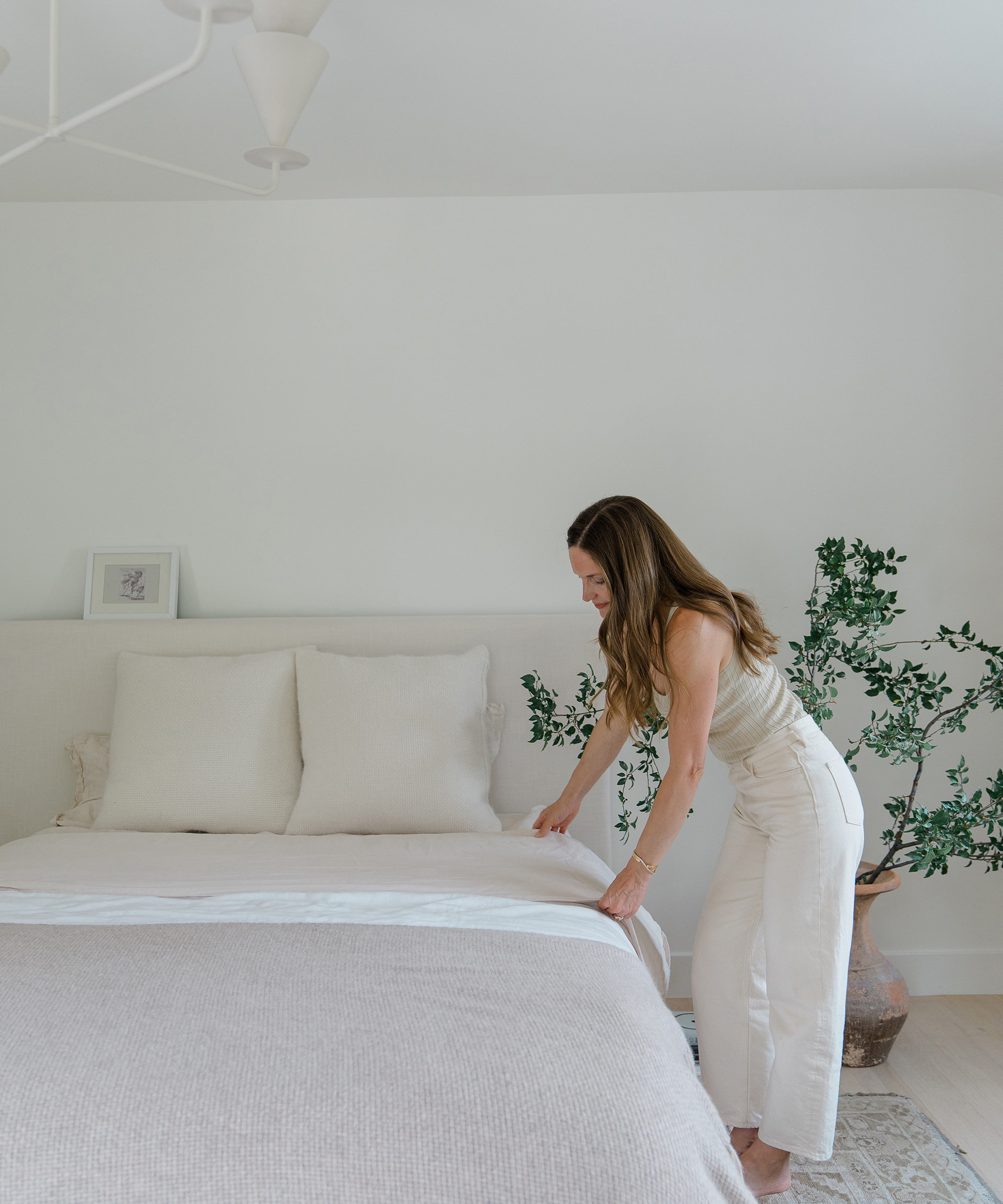 A woman with long brown hair in light-colored clothes is making a neatly arranged bed in a minimal, modern interior bedroom with white walls and a large potted plant.