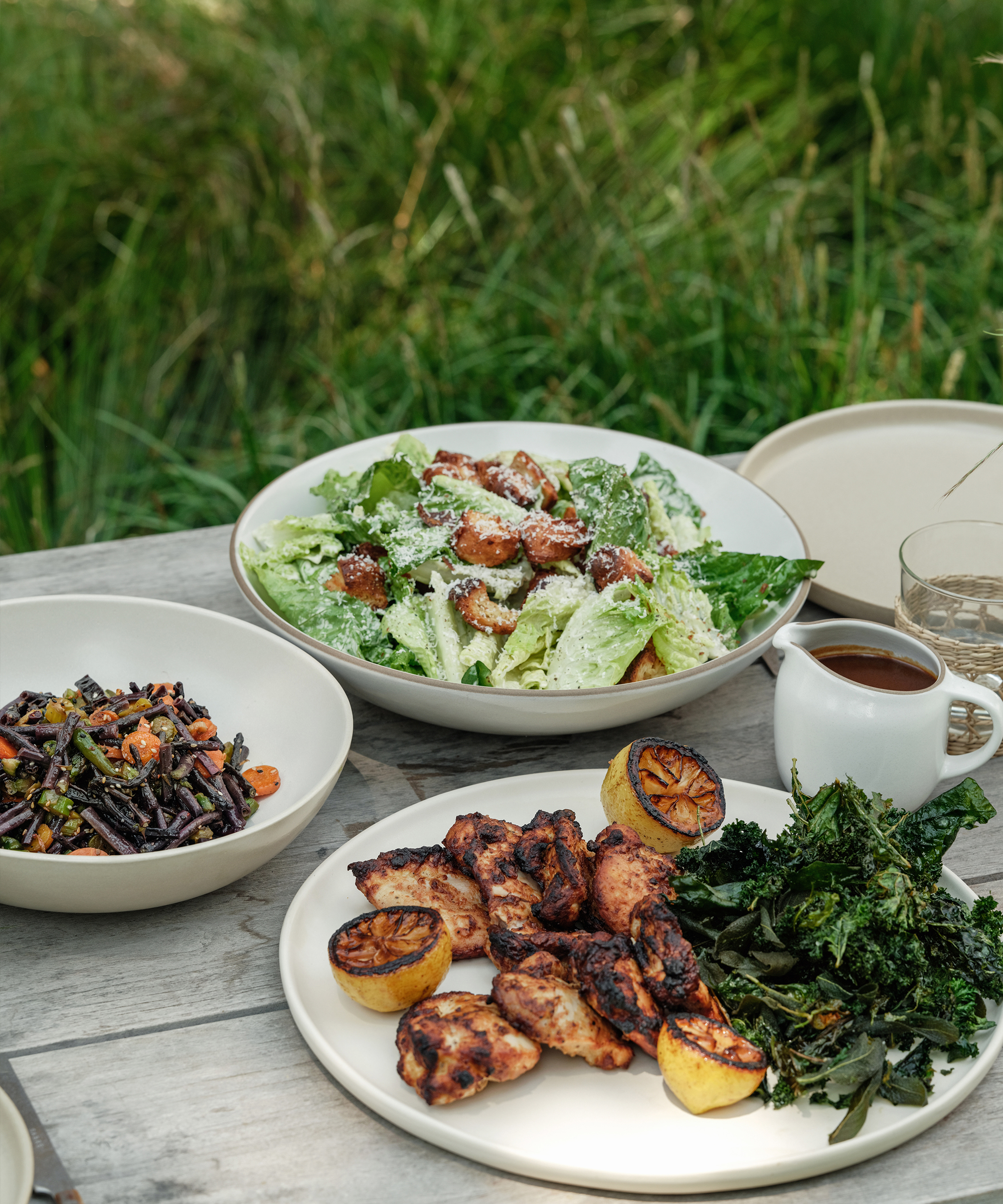 a bowl of ceasar salad and a plate or roasted check and roasted veggies on a wood outdoor dining table