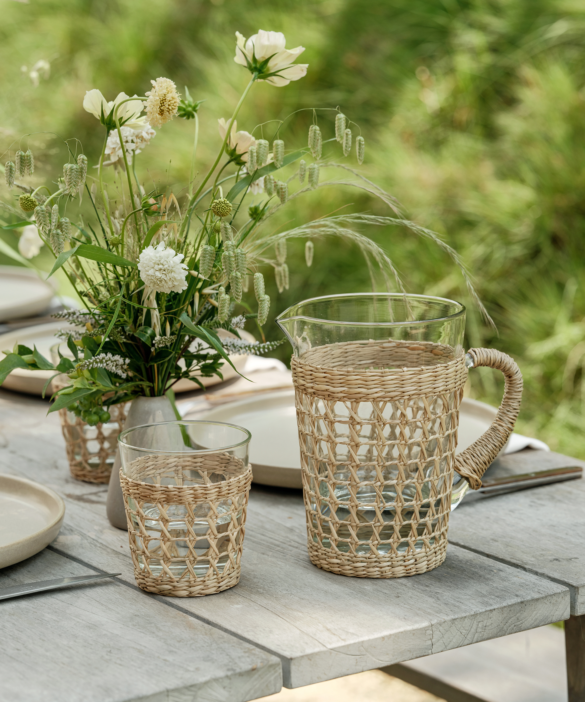A glass pitcher and tumbler with woven wicker covers sit on an outdoor wooden table next to a vase of white wildflowers—echoing the fresh, inviting vibes of Sqirl—with plates and napkins set for a meal. Lush greenery fills the background.
