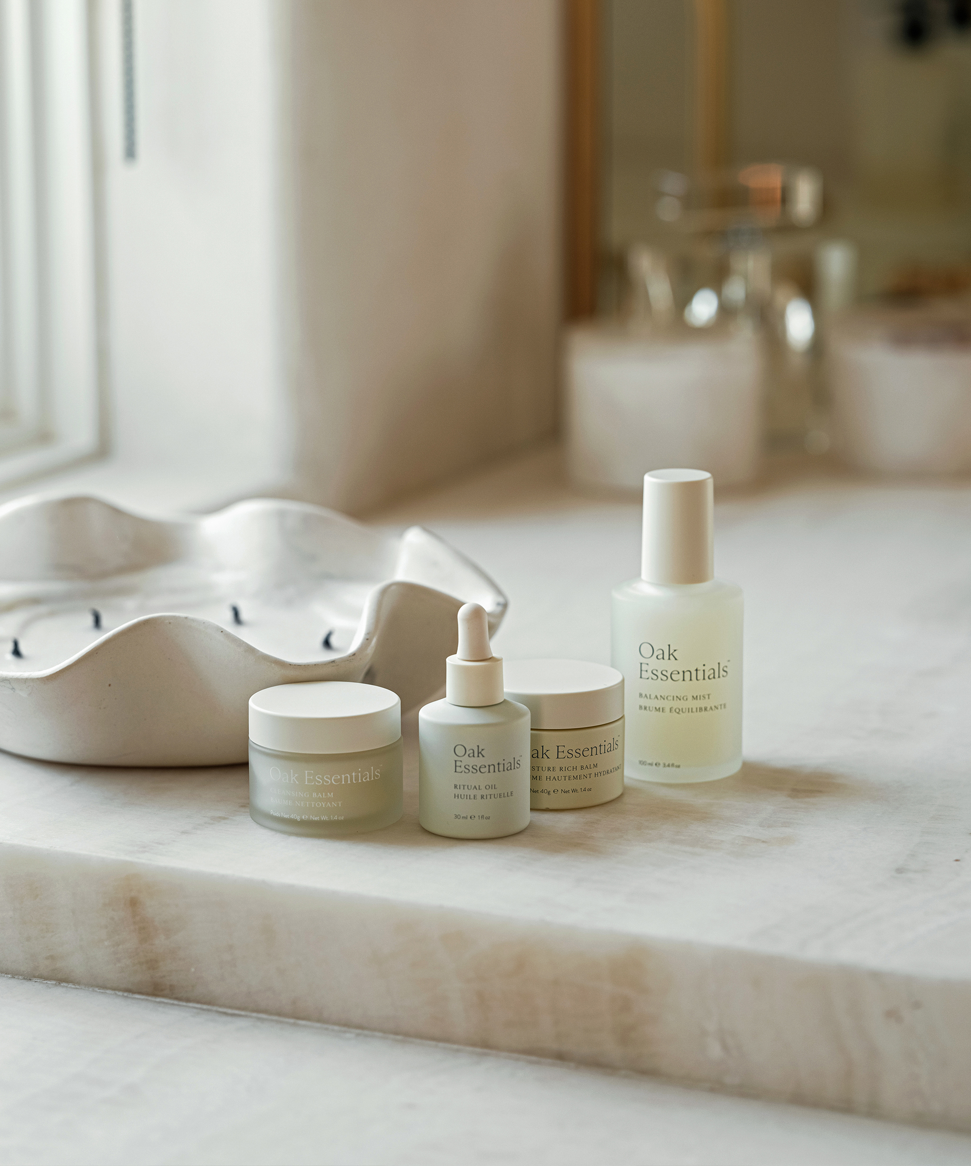 Four Oak Essentials skincare products, loved by Tracy Tutor, are arranged neatly on a marble countertop near a window and a decorative white candle.