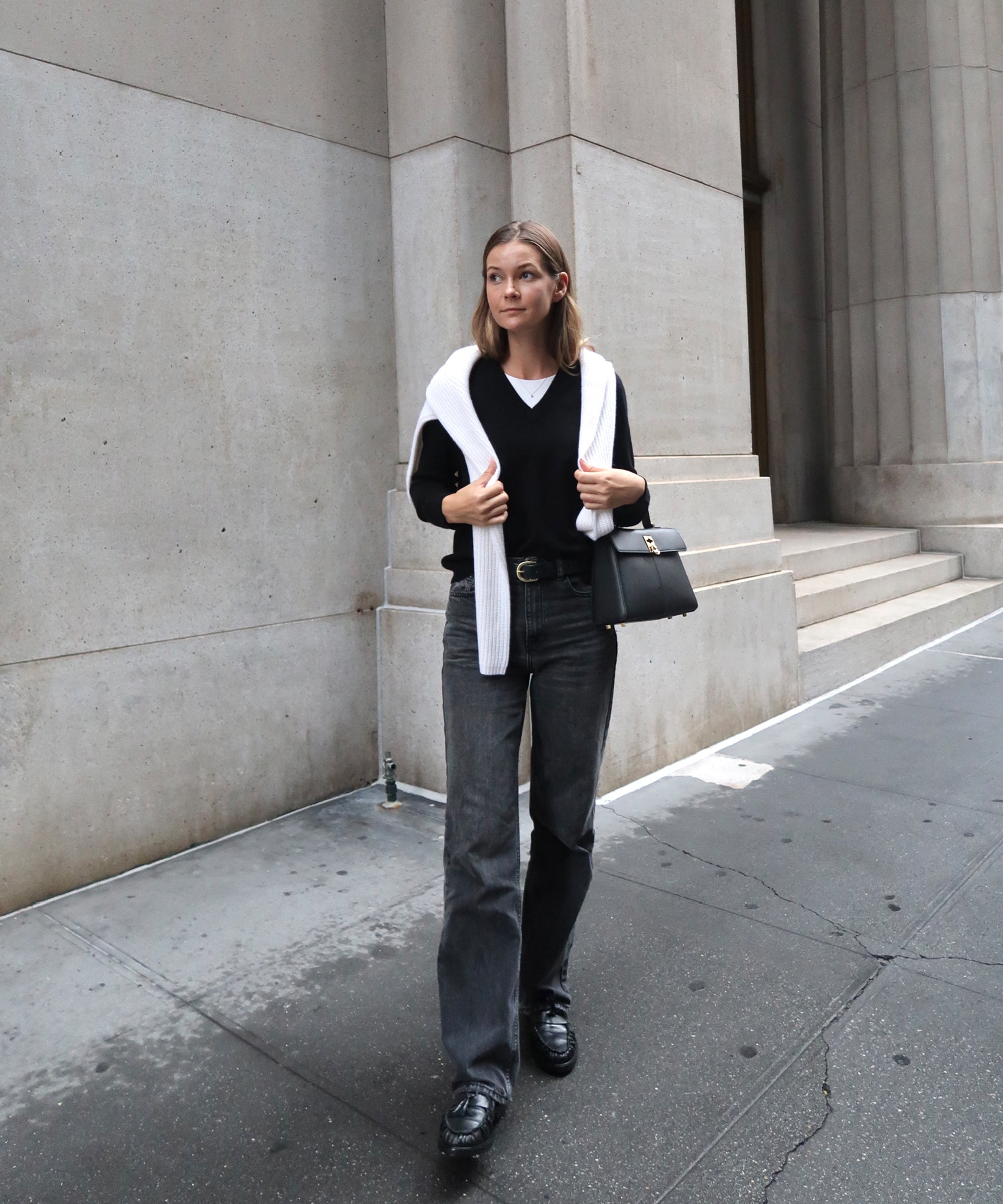 kristel is in a black v neck sweater with an ivory sweater layered and black jeans standing in front of a concrete wall