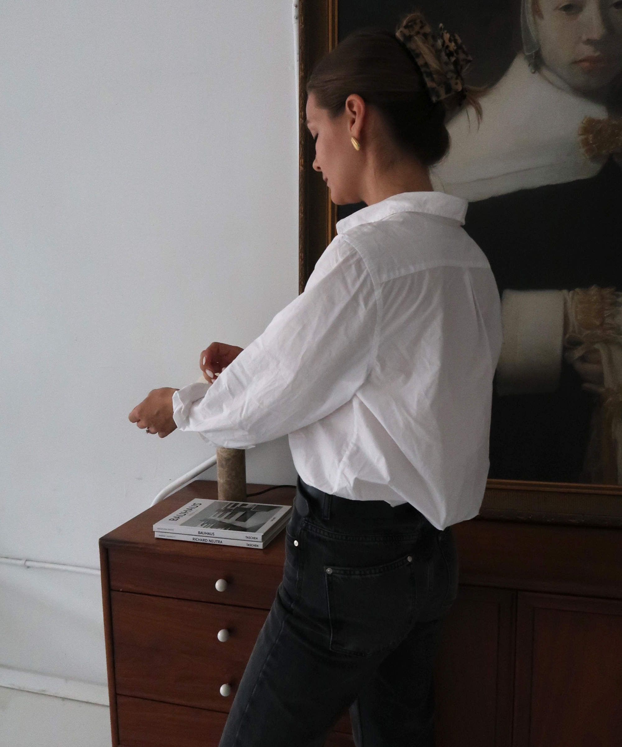 A woman with hair in a clip, wearing a white button-down shirt and black jeans—classic fall outfits—stands by a wooden dresser adjusting her sleeve. Books and a cylindrical object rest on the dresser, with a portrait painting in the background.