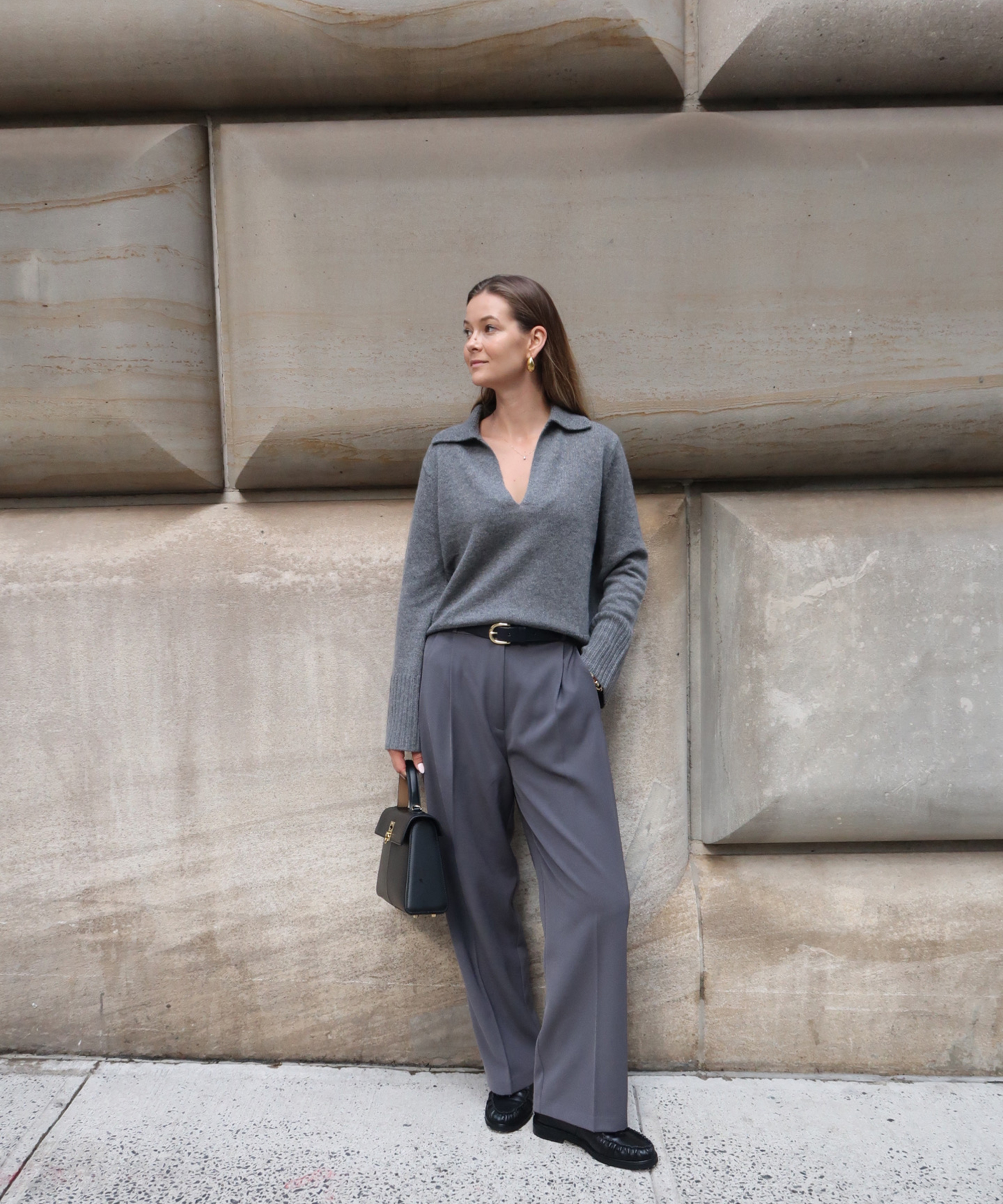 kristel standing in front of a concrete wall in a grey frances polo sweater and grey trousers with black loafers