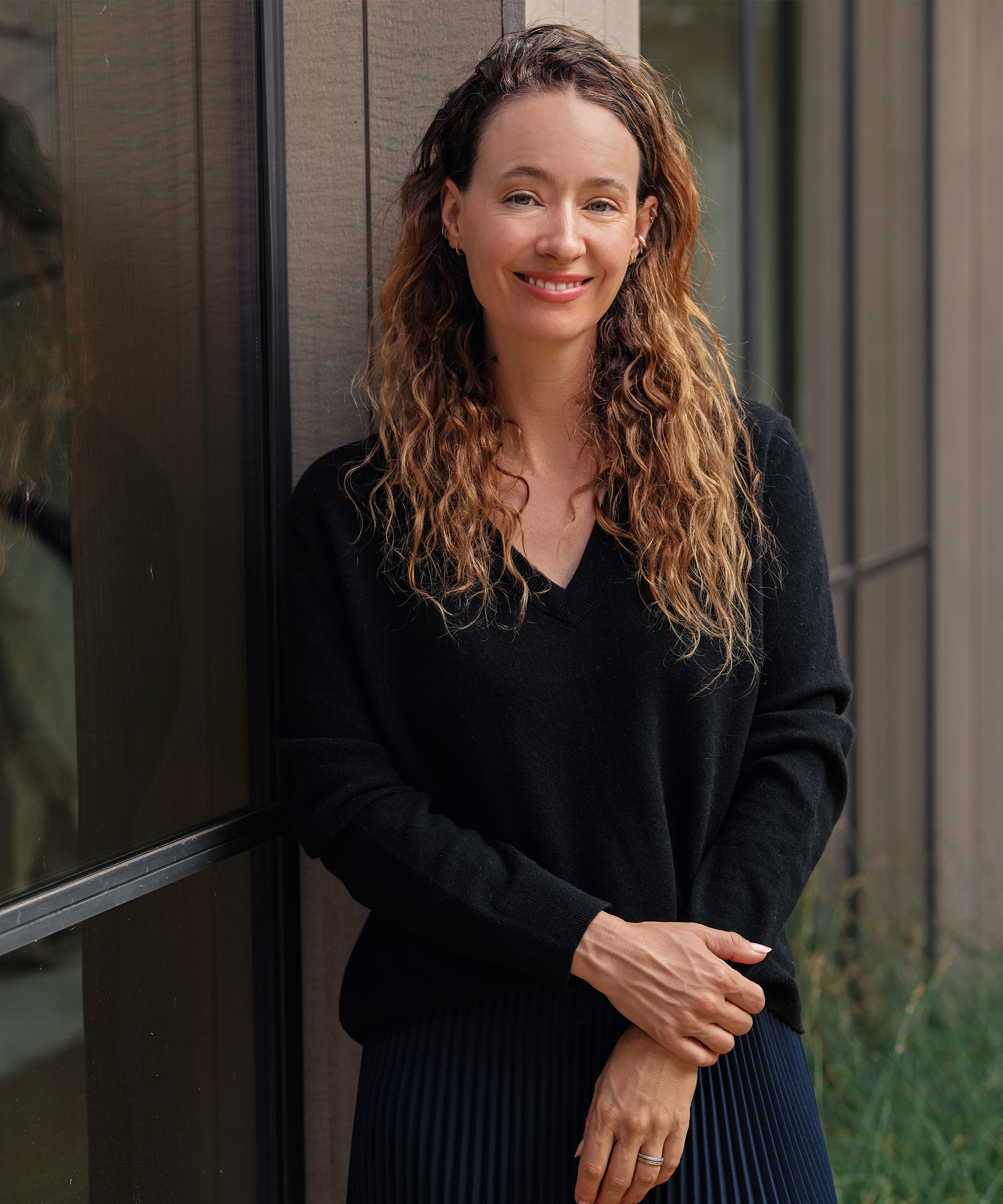 A woman with long wavy brown hair and a black sweater smiles while standing with arms crossed in front of a window and wooden wall, showcasing a cozy fall outfit.