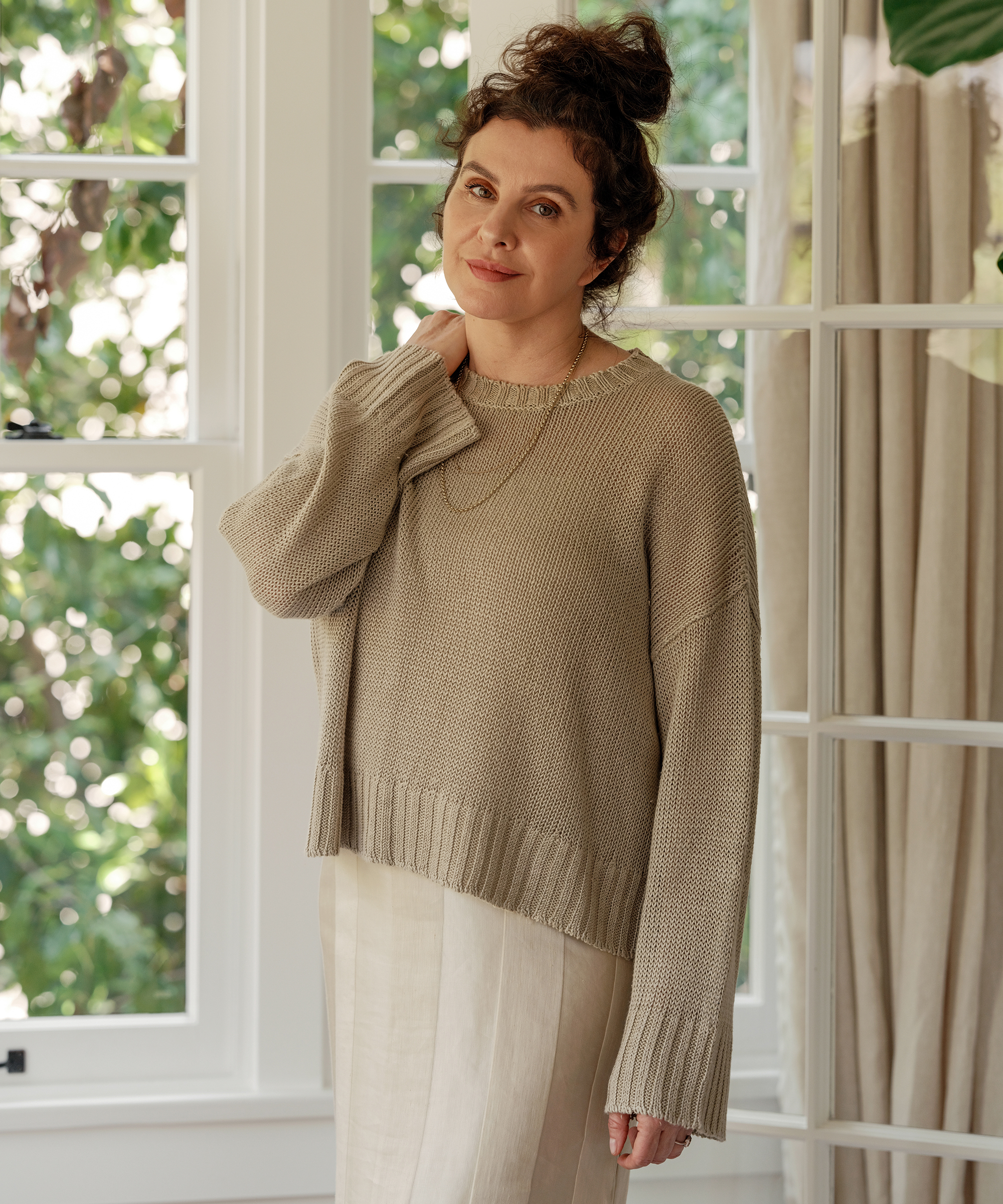 A woman with curly hair in a loose bun, wearing a beige knit sweater and light skirt, stands by a window in Hancock Park with greenery outside. She smiles softly and lightly touches her collar, sunlight streaming in behind her.
