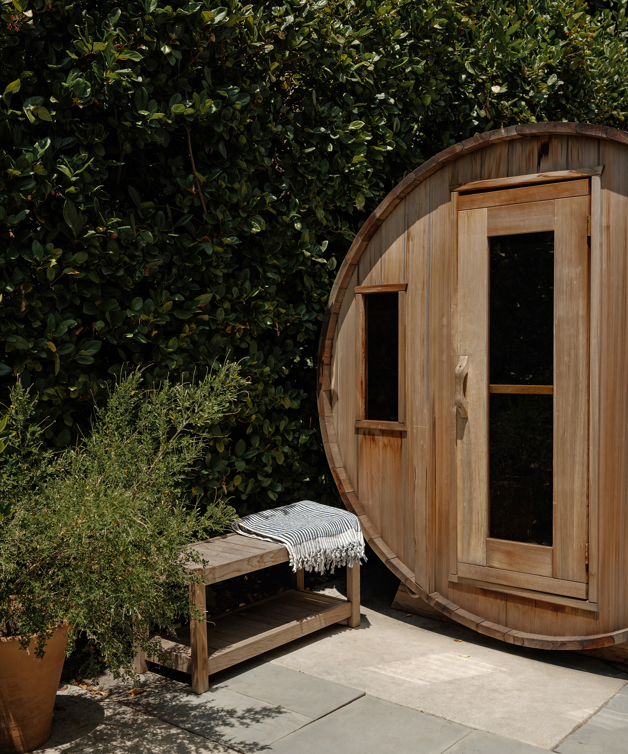 A round wooden sauna sits beside a leafy green hedge in Hancock Park. In front, a small wooden bench with a folded striped towel and a potted plant rests on a stone patio bathed in bright sunlight.