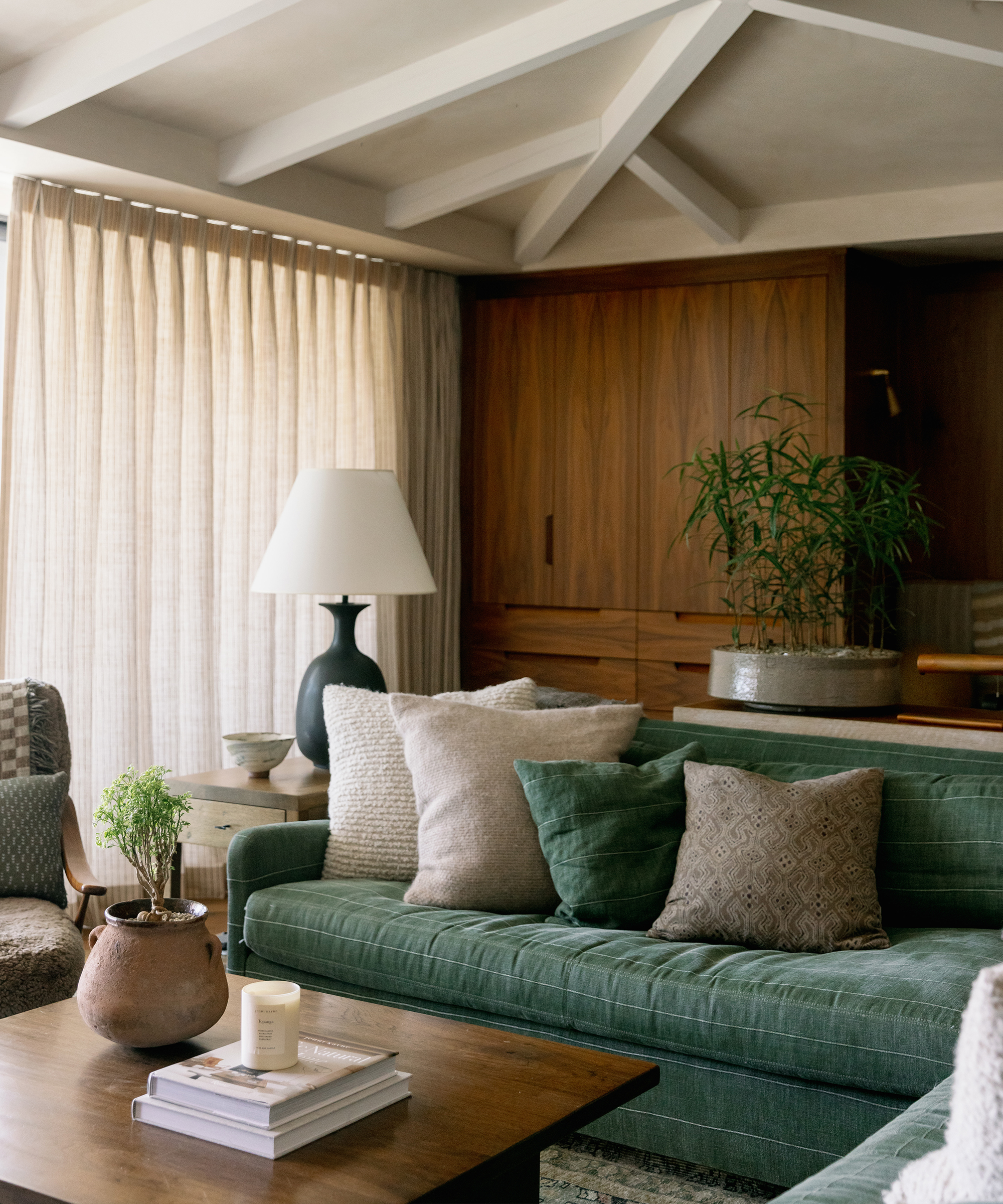 A cozy living room with an emerald bay green sofa, assorted pillows, a wooden coffee table with books and a potted plant, a table lamp, and houseplants. Light filters through beige curtains, creating a warm, inviting atmosphere.