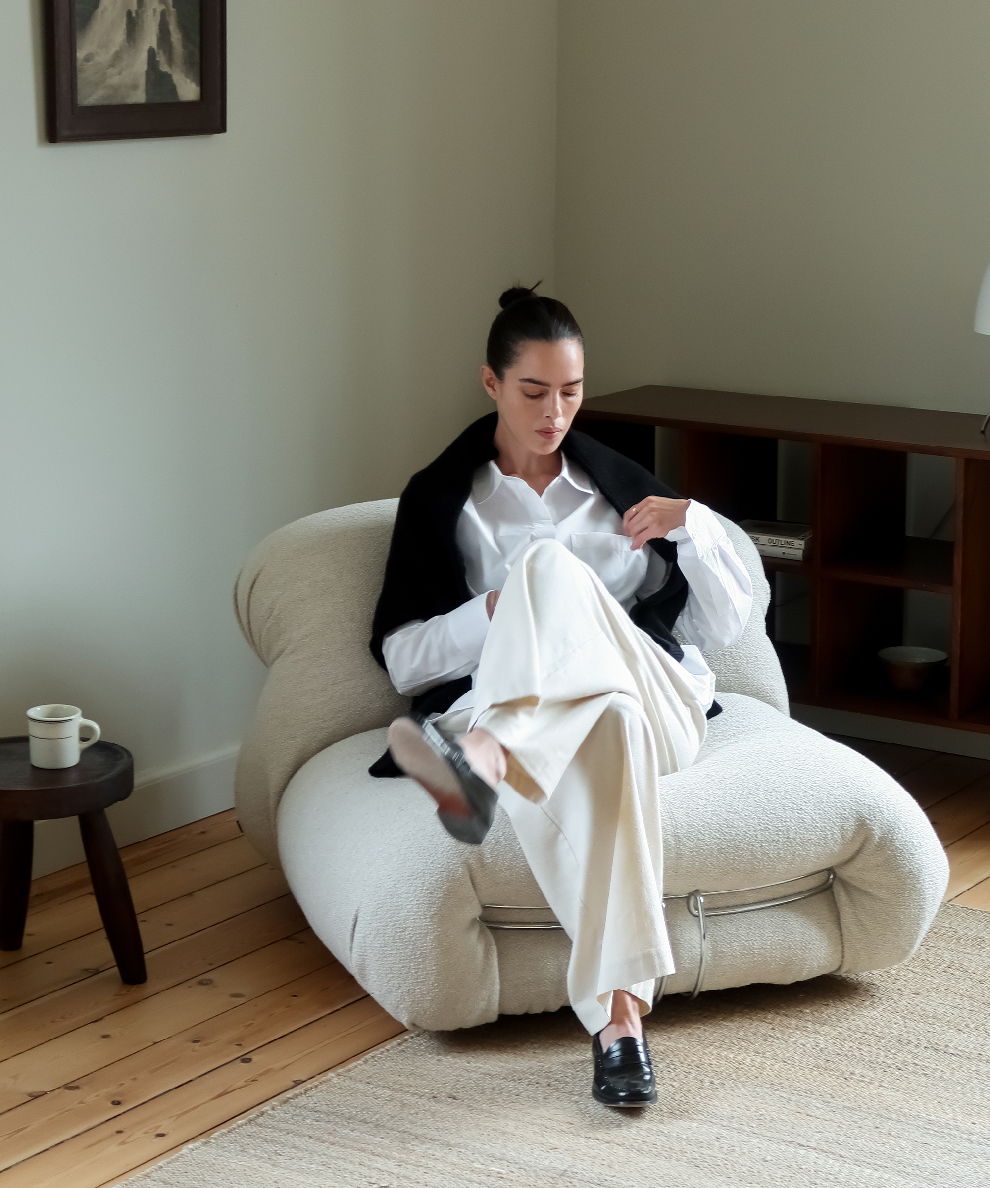 a woman sitting in a boucle accent chair in ivory trousers and a boyfriend shirt with a black cardigan over her shoulders