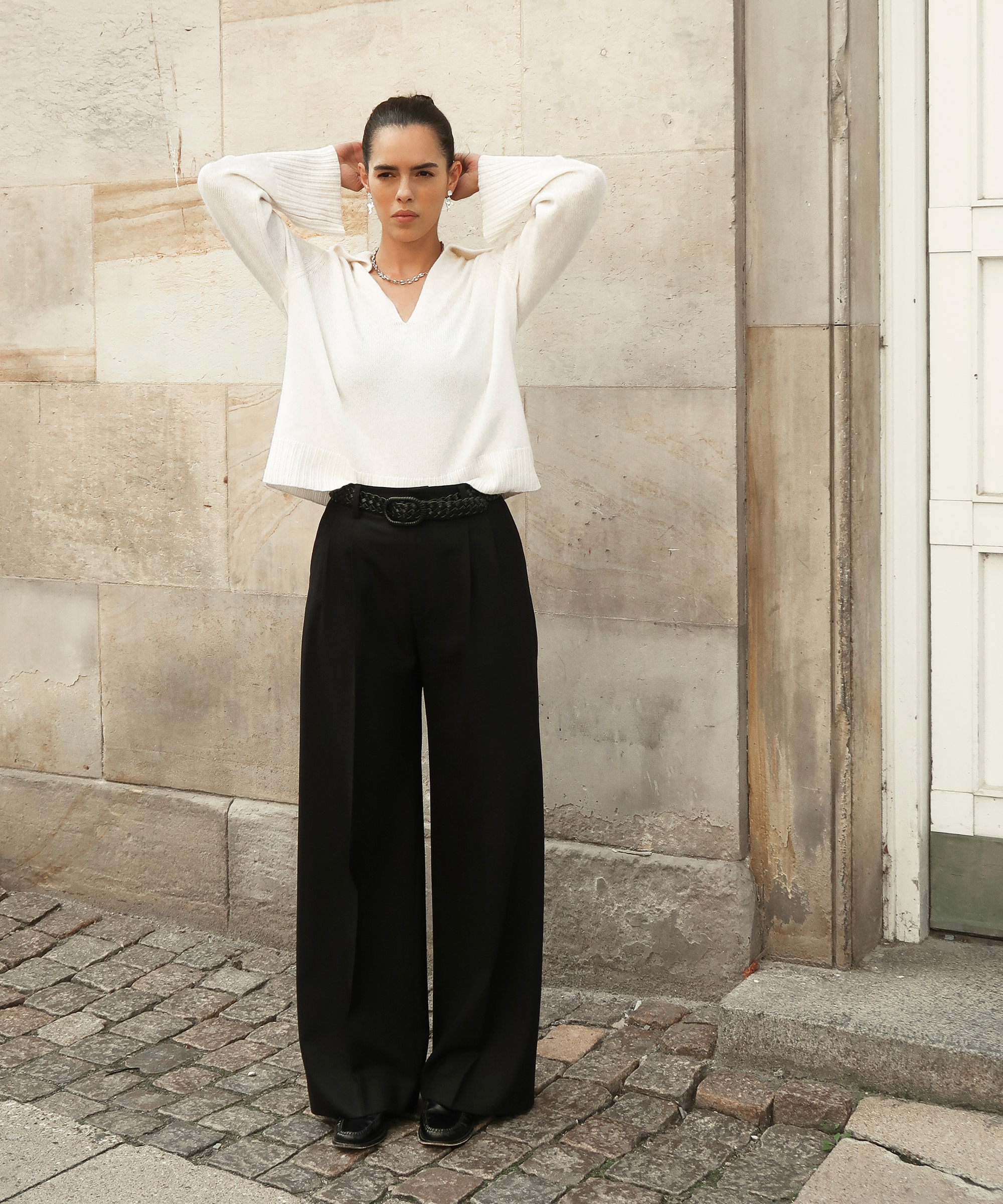 a woman in black trousers and an ivory cashmere frances polo standing in front of stone wall