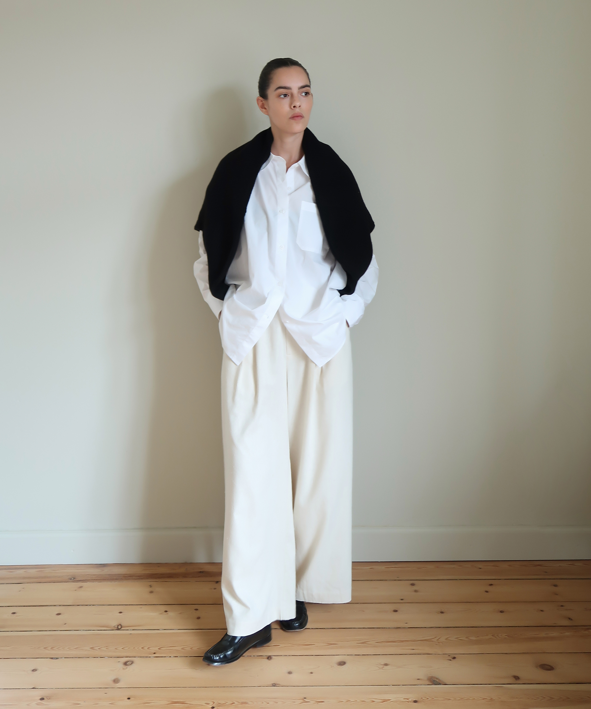 a woman in ivory trousers with a white button up shirt and a black cardigan standing in front of a wall