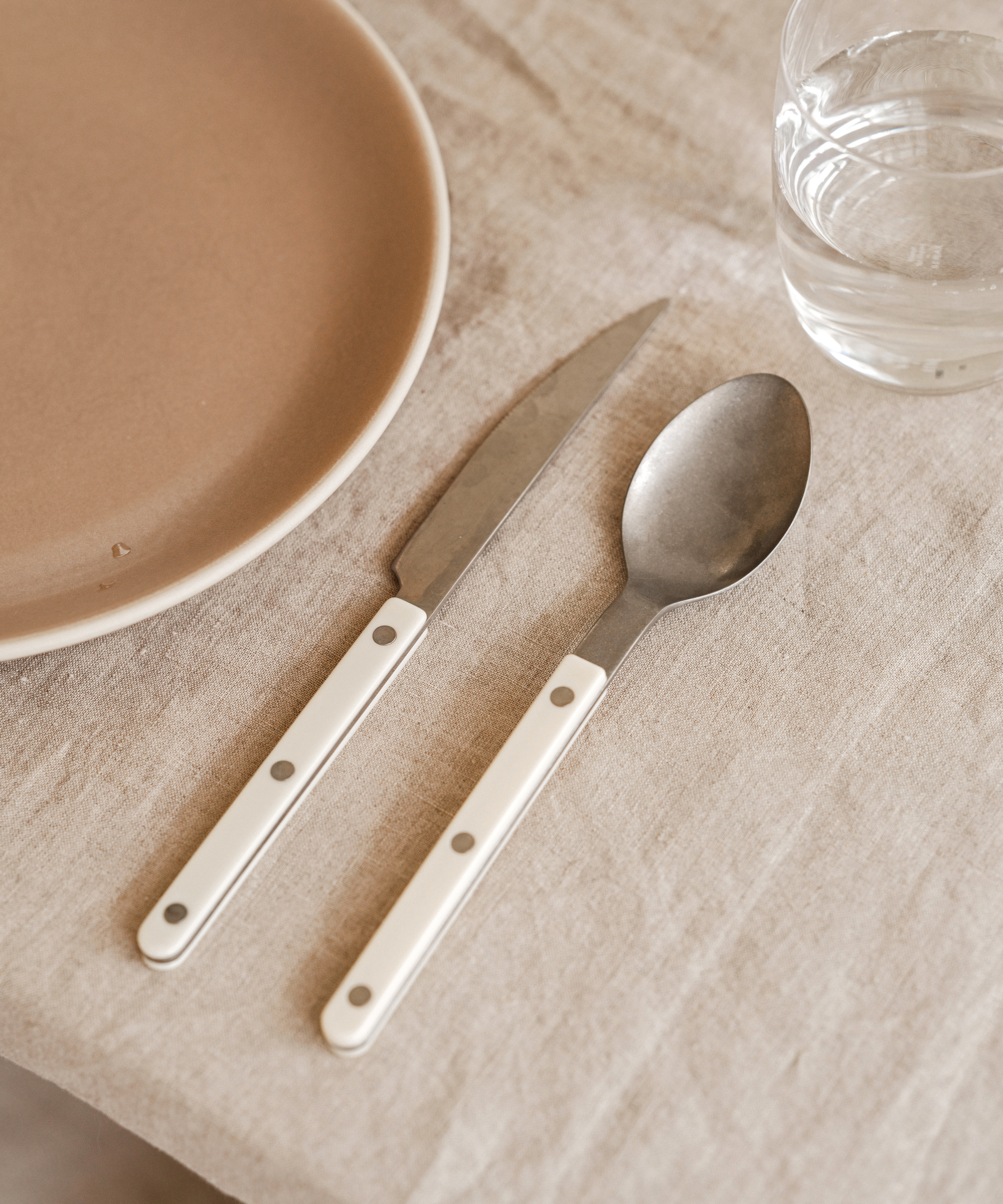 A beige plate, a knife, and a spoon with white handles are arranged on a light fabric tablecloth next to a clear glass of water—perfect for serving your favorite fall vegetarian recipe.
