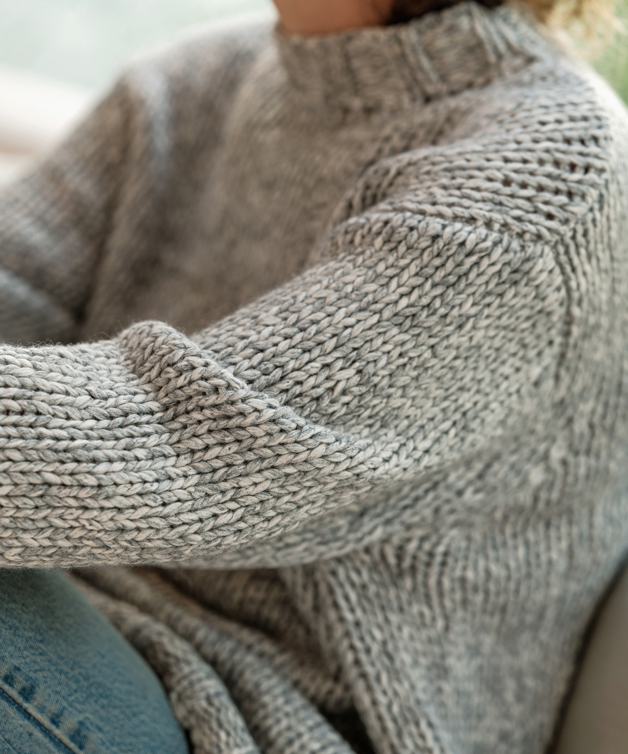 A person wearing a thick, light gray knitted sweater and blue jeans sits comfortably, embracing sweater weather. Their face is partially out of frame, while the texture and pattern of the sweater are clearly visible.