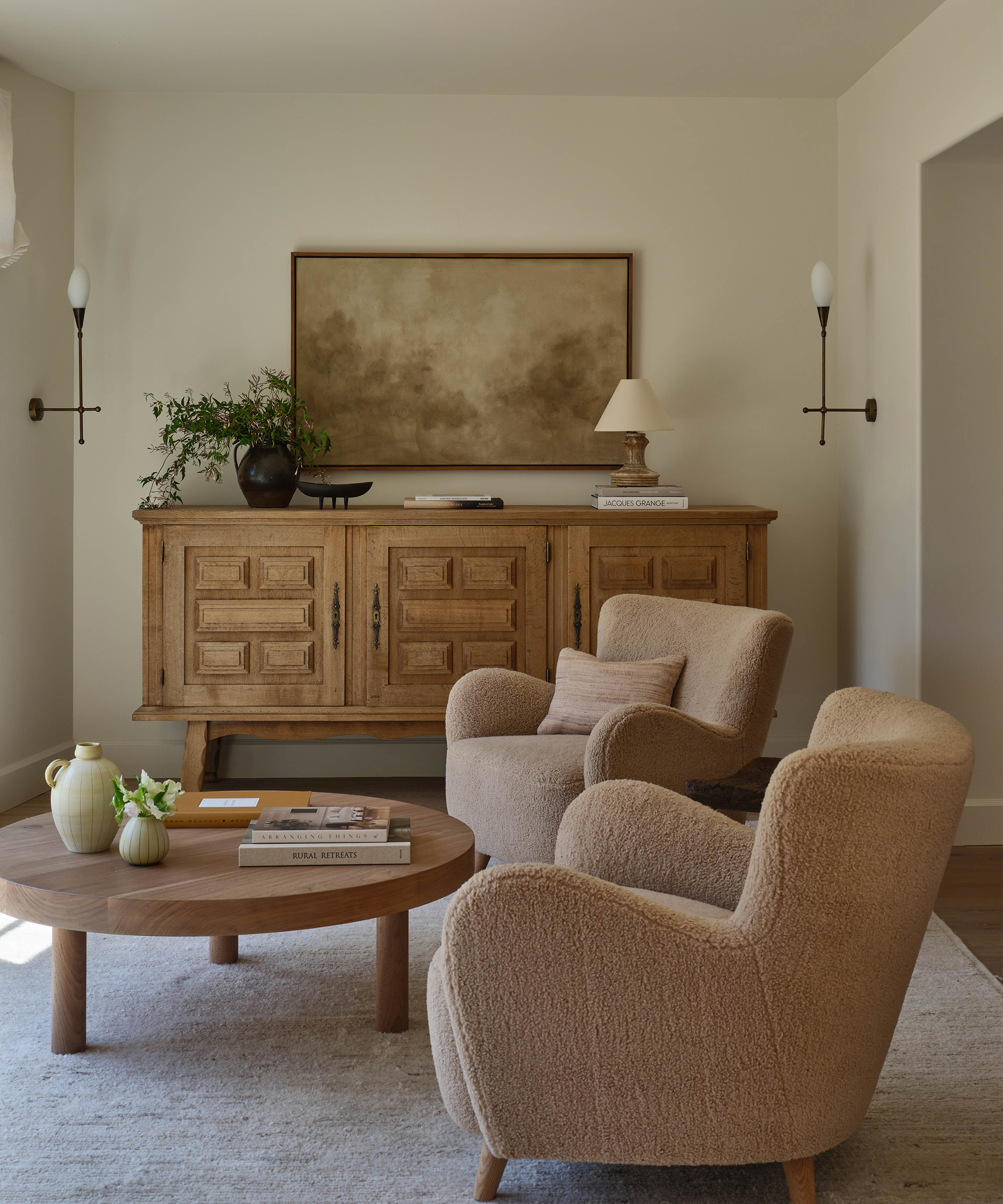 two sherpa taupe accent chairs in front of a round wooden coffee table next to an antique sideboard with a painting hung above