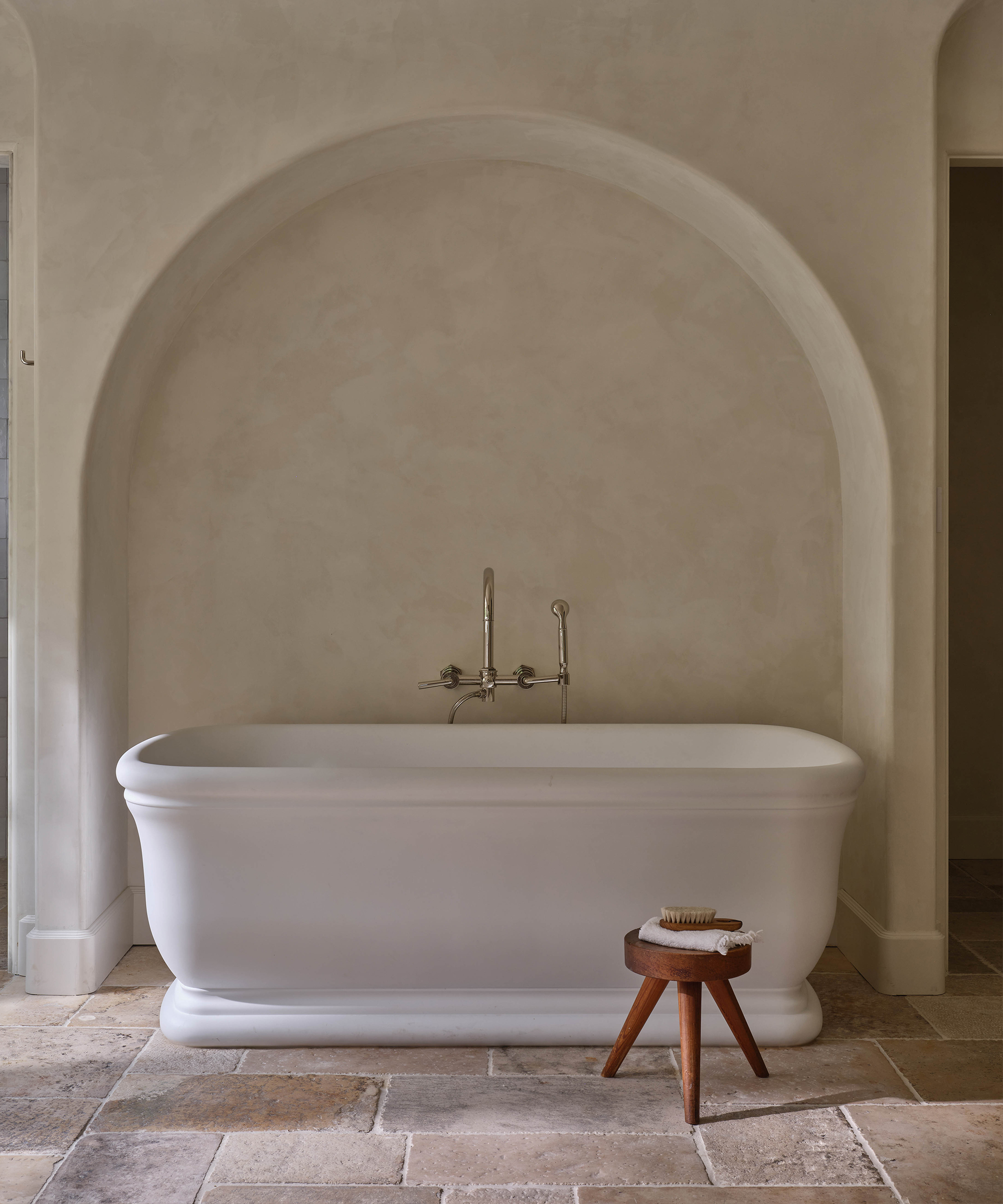 A white freestanding bathtub sits in front of an arched alcove with wall-mounted fixtures, revealing a stunning before and after transformation. A small wooden stool with a brush rests on stone tile flooring in this minimalist, serene space.