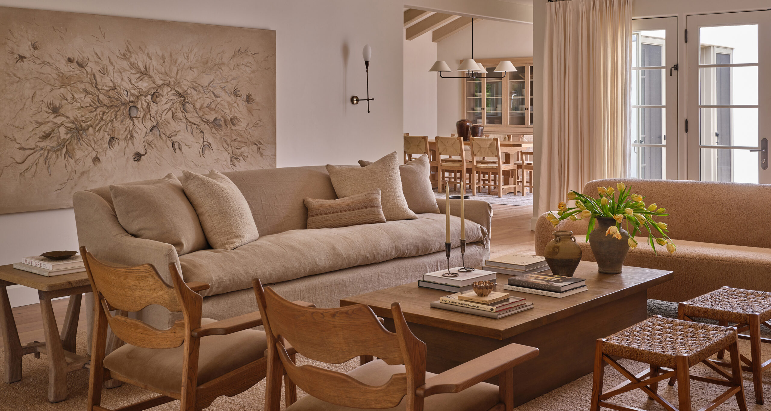 A cozy living room with neutral tones features a beige sofa, wooden chairs, and woven stools. Sunlight streams through glass doors, highlighting the inviting space—perfect for before and after photos that capture every design detail.