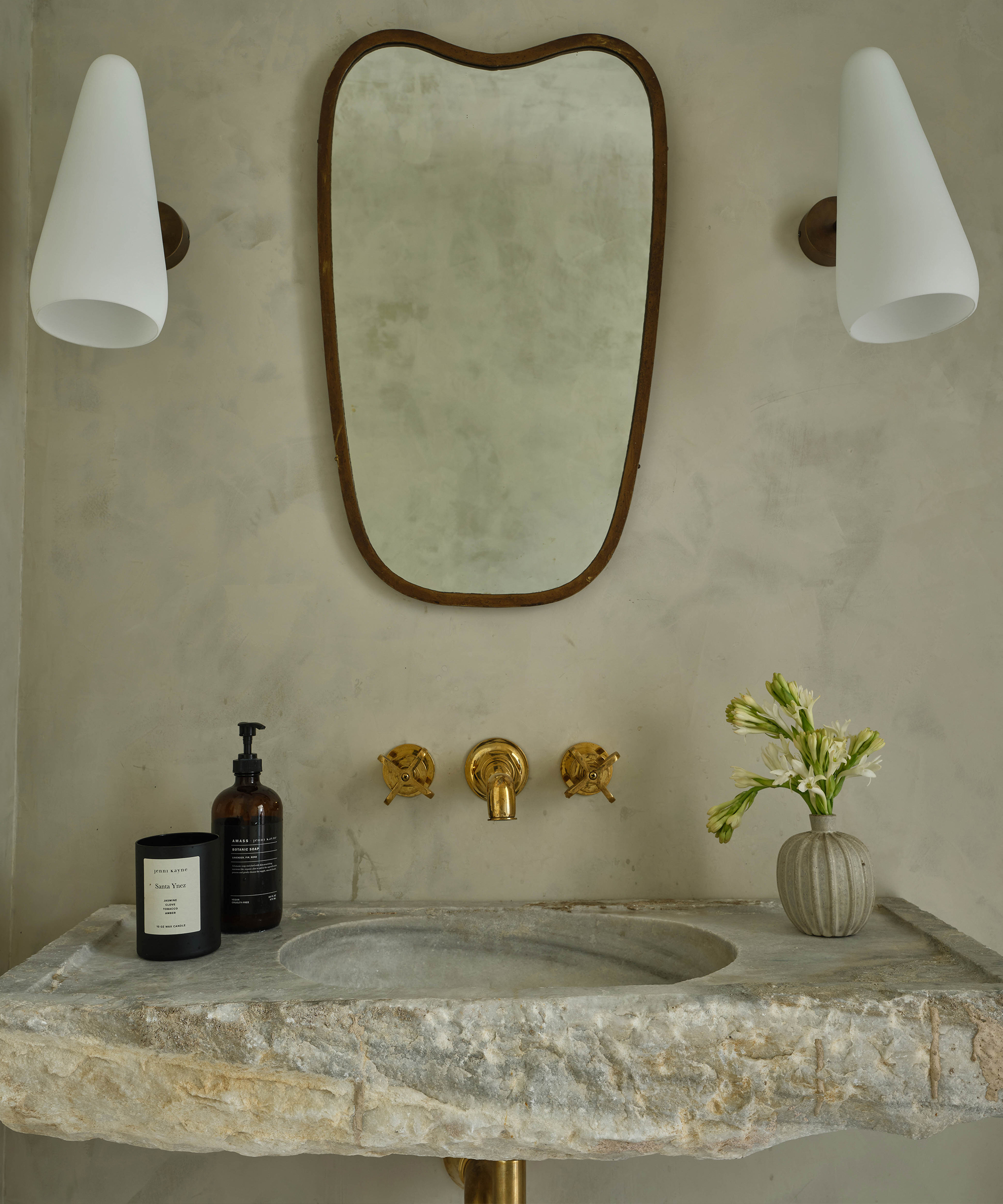 A rustic bathroom sink with a stone countertop, brass faucet fixtures, and a small vase with green flowers creates the perfect before and after transformation, complemented by an irregular mirror flanked by white sconces on a textured beige wall.