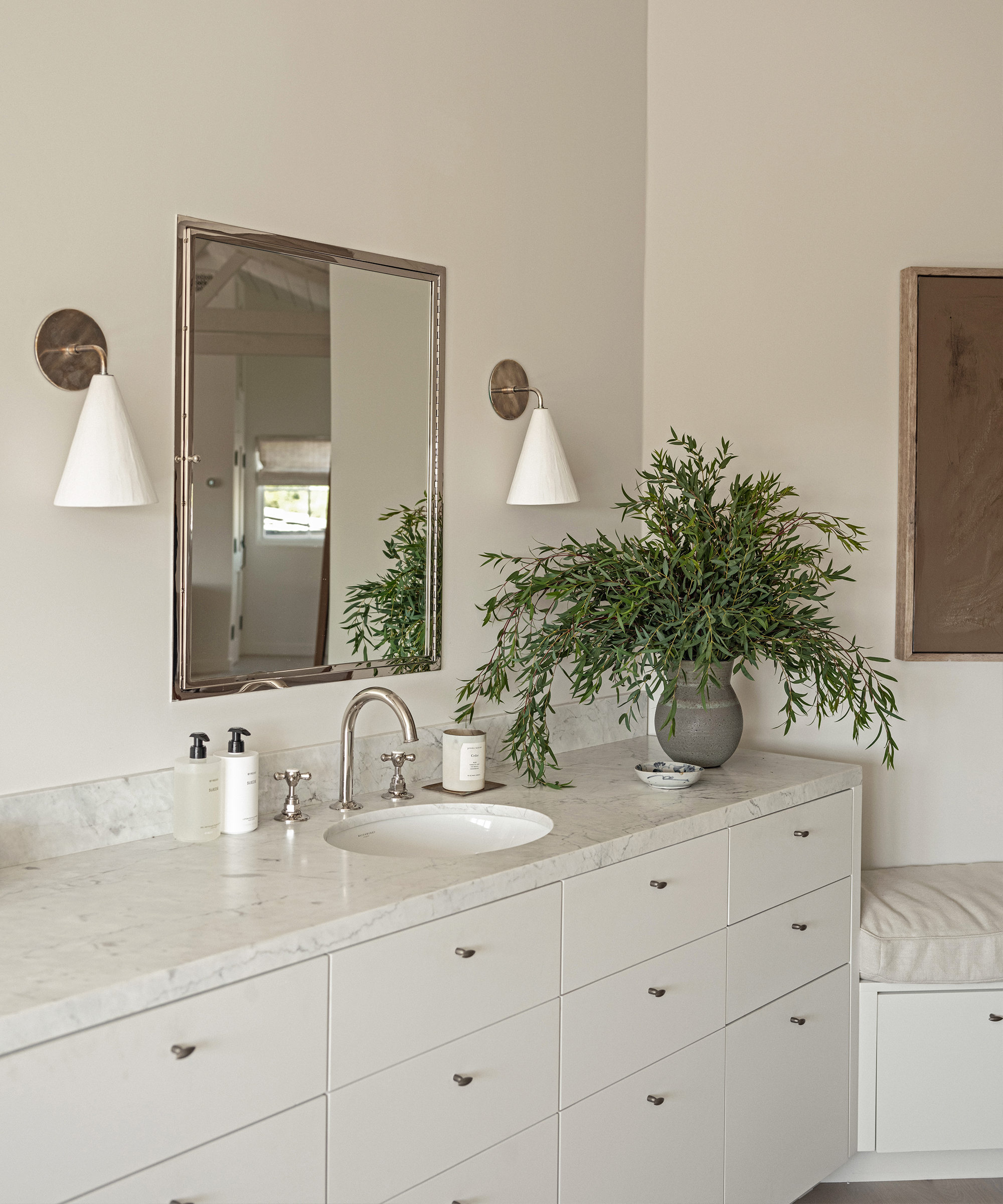 A modern bathroom vanity with white drawers, a marble countertop, a silver-framed mirror, two wall sconces, soap dispensers, and a large vase with green leafy branches—showcasing the neat method in neutral, minimalist decor.