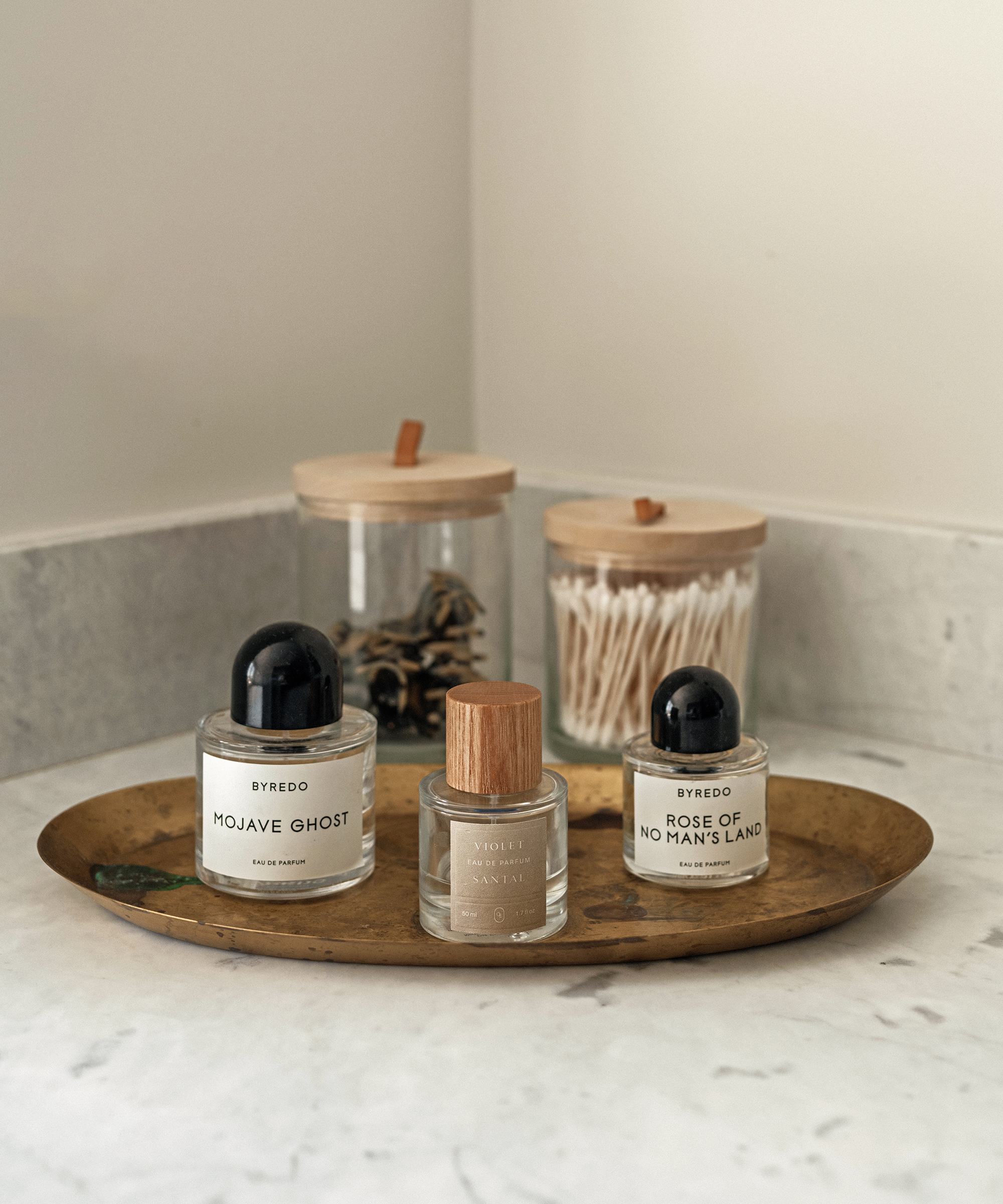 Three perfume bottles sit on a brass tray on a marble countertop, with two glass jars—one filled with cotton swabs and one with miscellaneous items—in the background, showcasing a neat method for keeping the setting minimal and clean.