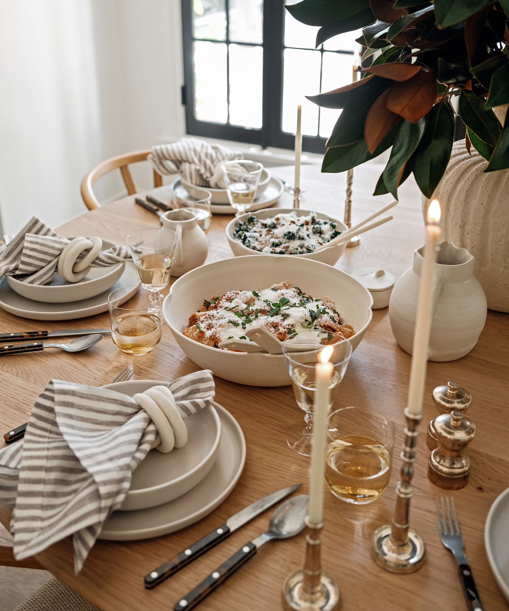 a dining table with a large bowl of pasta and place settings with ivory plates and striped napkins