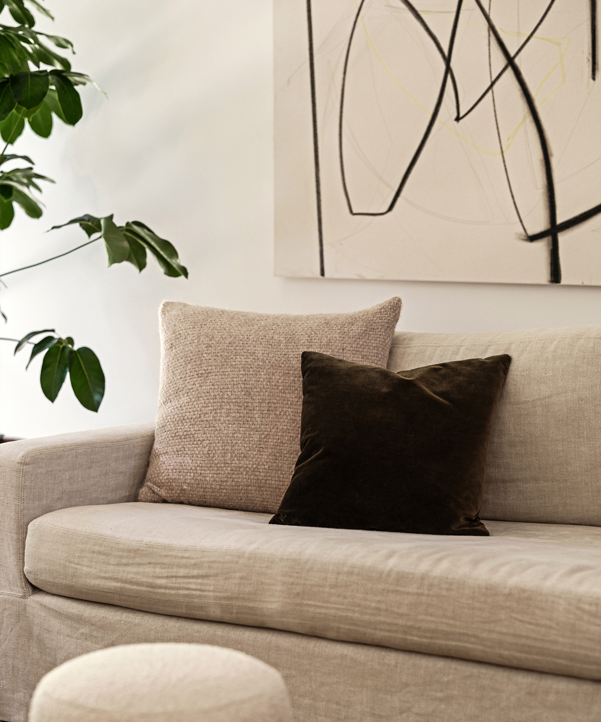 A beige Brentwood sofa with two throw pillows, one light textured and one dark velvet, sits beneath an abstract black and yellow line art painting. A leafy green plant is beside the sofa.