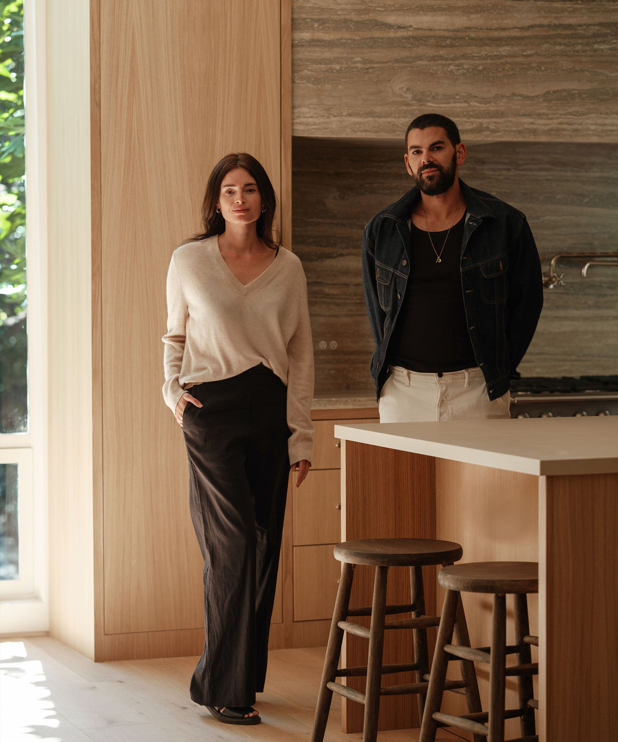 A woman and a man stand in a modern Brentwood kitchen with light wood cabinets and stone walls. The woman wears a beige sweater and dark pants, while the man wears a black shirt, denim jacket, and light pants. Two stools are at the island.