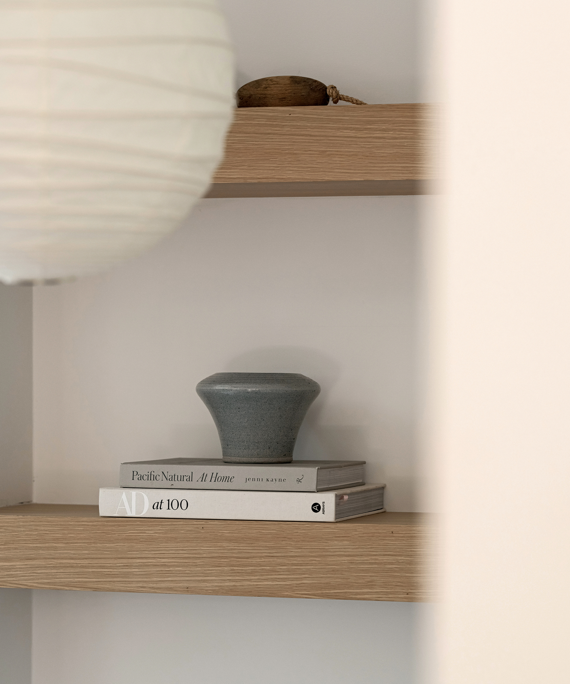 A minimalist wooden shelf in a Brentwood-inspired space holds a gray ceramic vase and two stacked books, Pacific Natural: At Home and AD at 100. Part of a white paper lampshade is visible on the left.