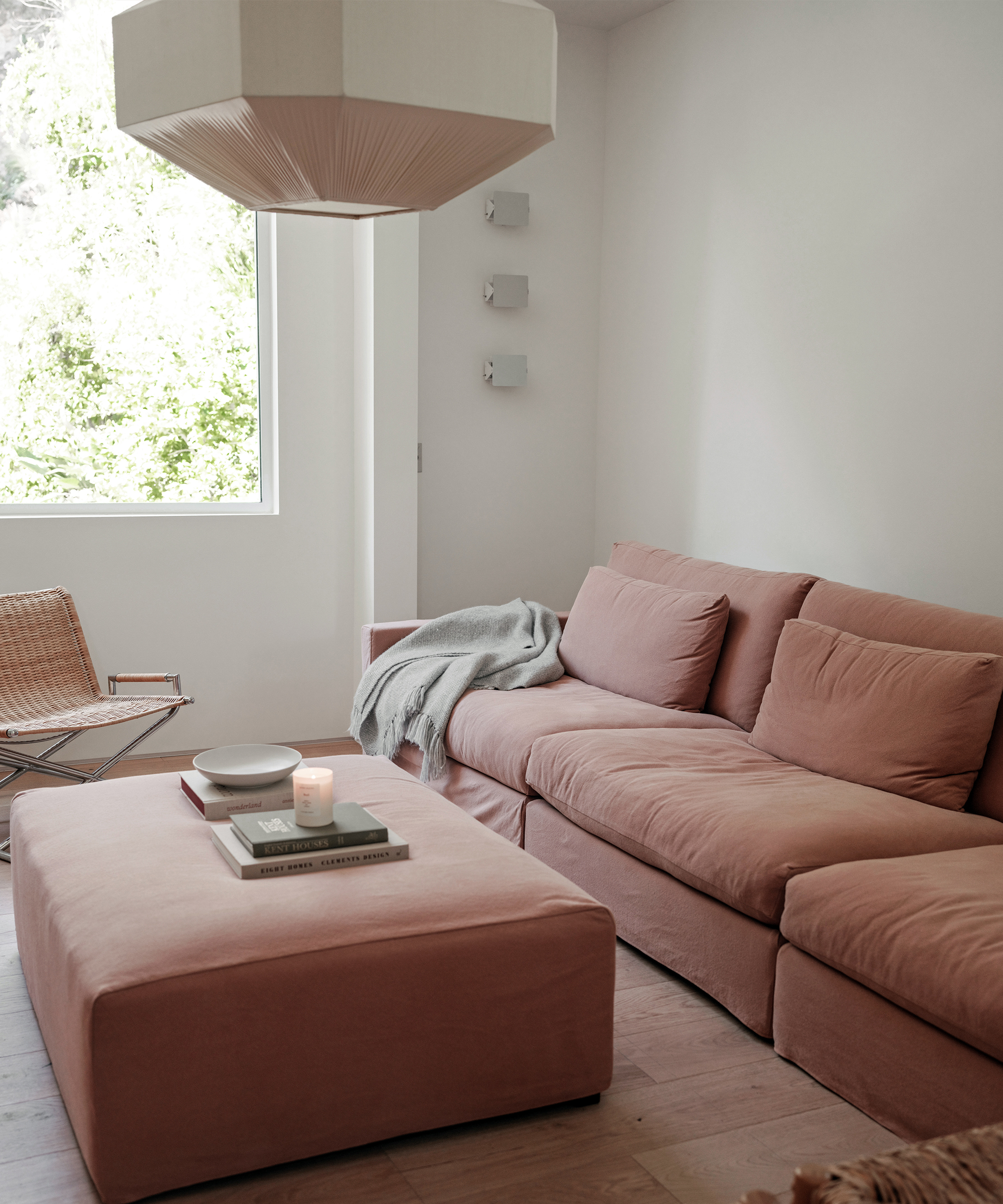 A cozy Brentwood living room with a blush pink sofa, matching ottoman, and light wooden floor. A candle, books, and bowl sit on the ottoman. Soft natural light enters through a window, with a modern pendant light above.