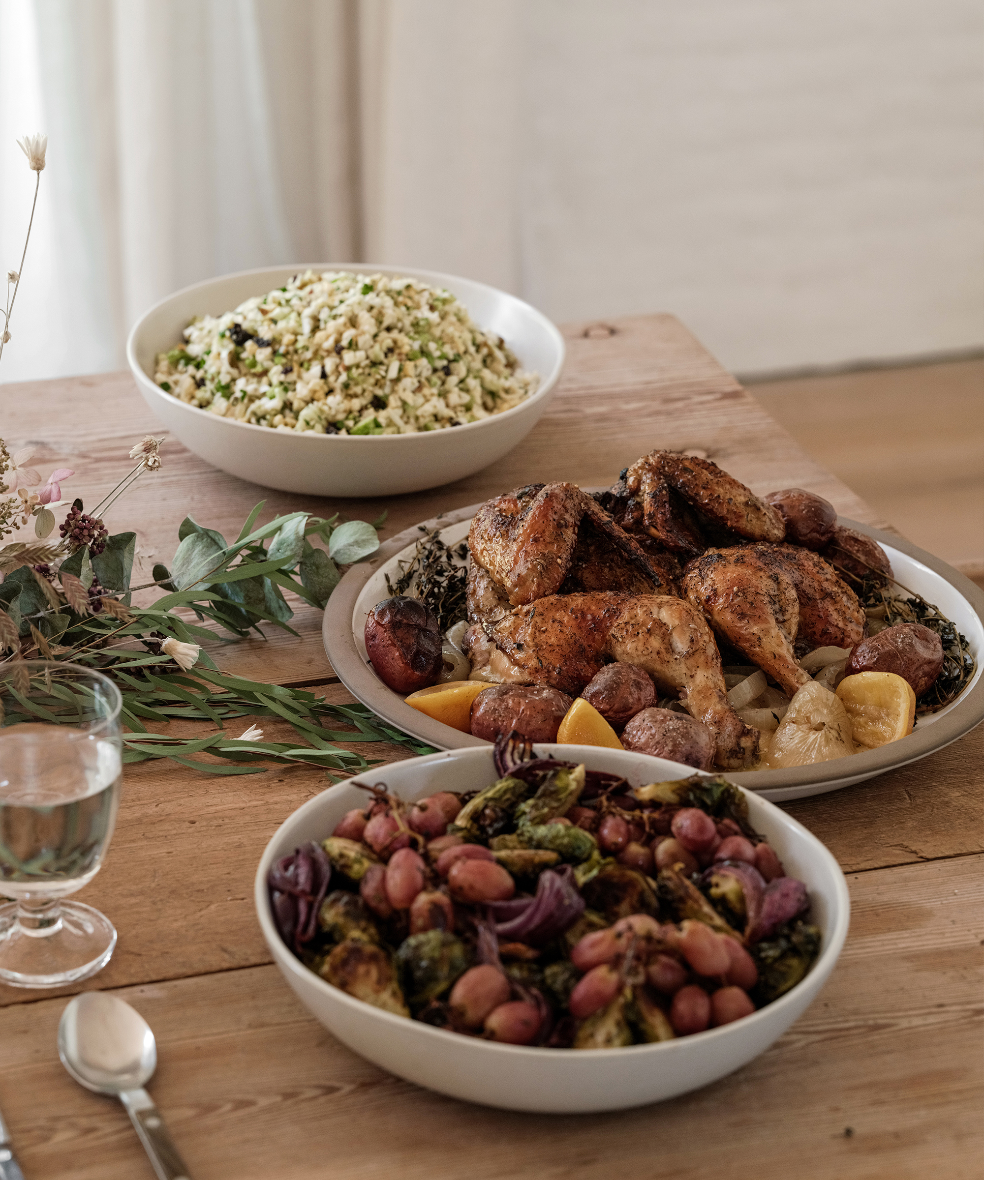 A wooden table set for a cozy fall dinner: three bowls—one with grilled chicken, potatoes, lemon, and herbs; one with couscous salad; and another with roasted vegetables and grapes. A glass of water and cutlery sit beside the dishes.