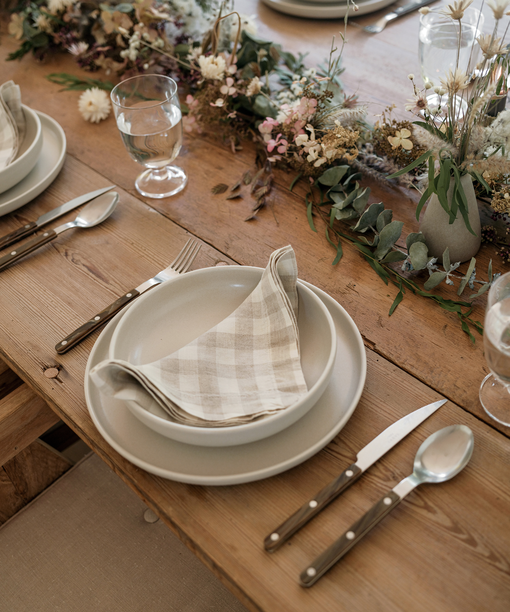 A rustic wooden table set for a cozy fall dinner features beige plates, checkered cloth napkins, silverware, and clear water glasses. A floral arrangement with dried flowers and greenery runs along the center.
