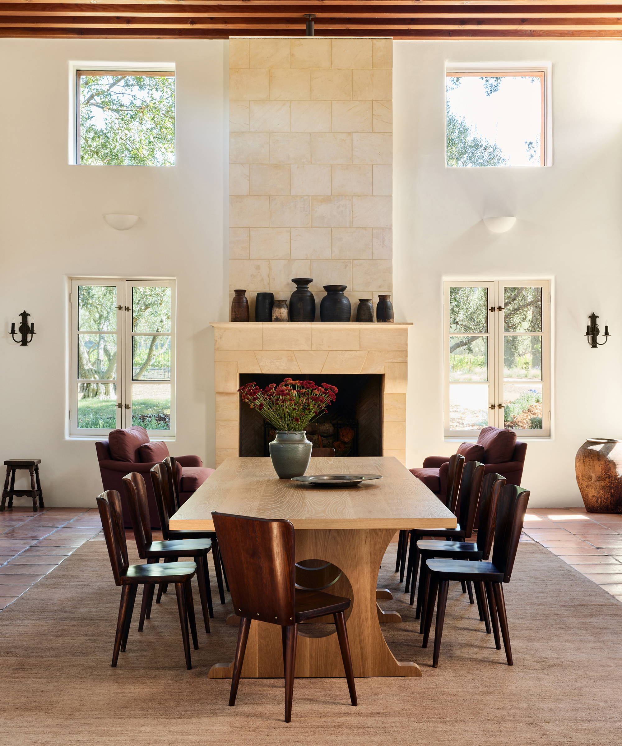 a dining room with dark wood dining chairs and a light oak dining table in front of a stone fireplace with vintage vases