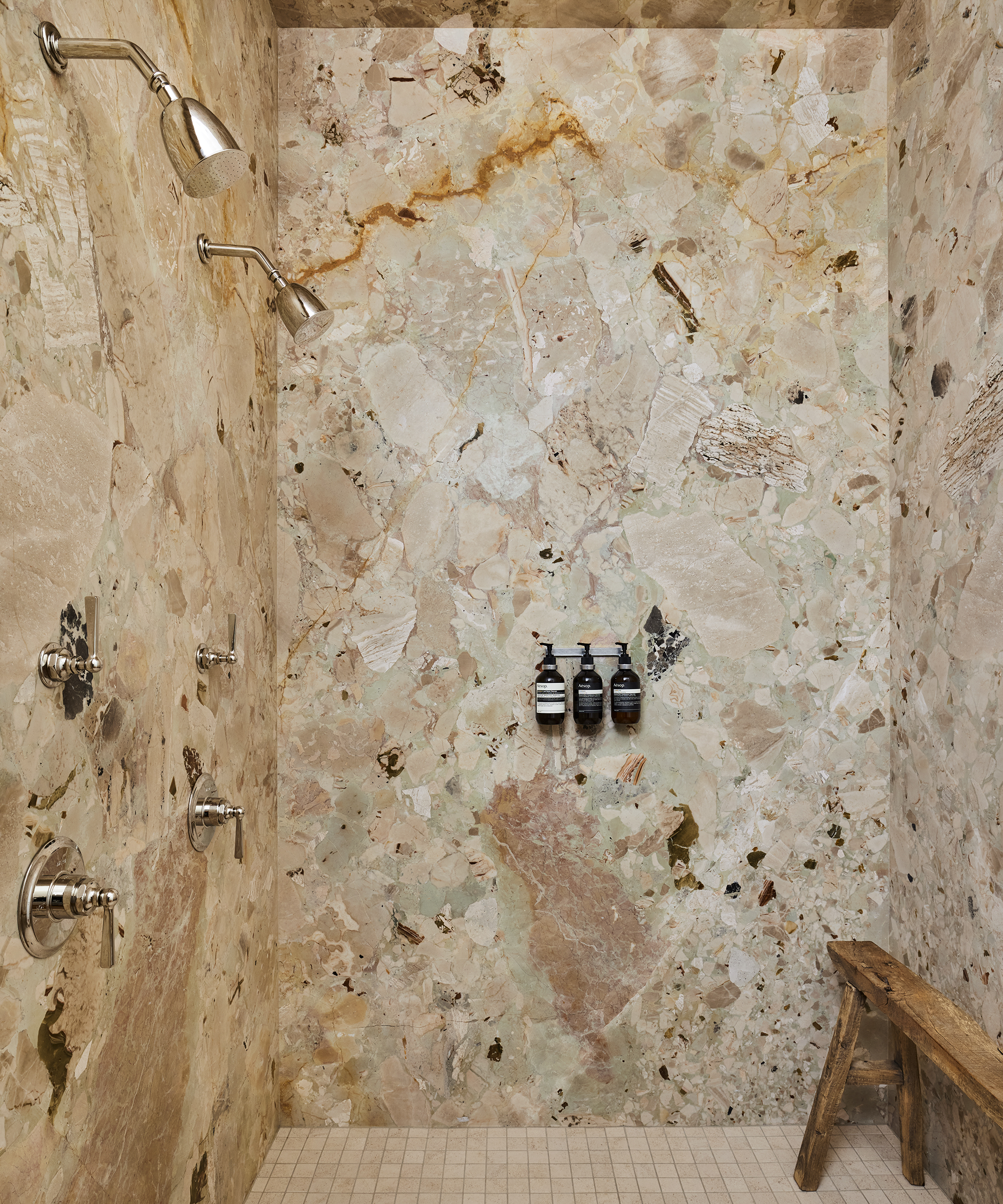A luxurious Corinne Mathern-inspired shower with marble-patterned walls, two showerheads, three wall-mounted controls, dark pump bottles, and a rustic wooden bench on a light tiled floor.