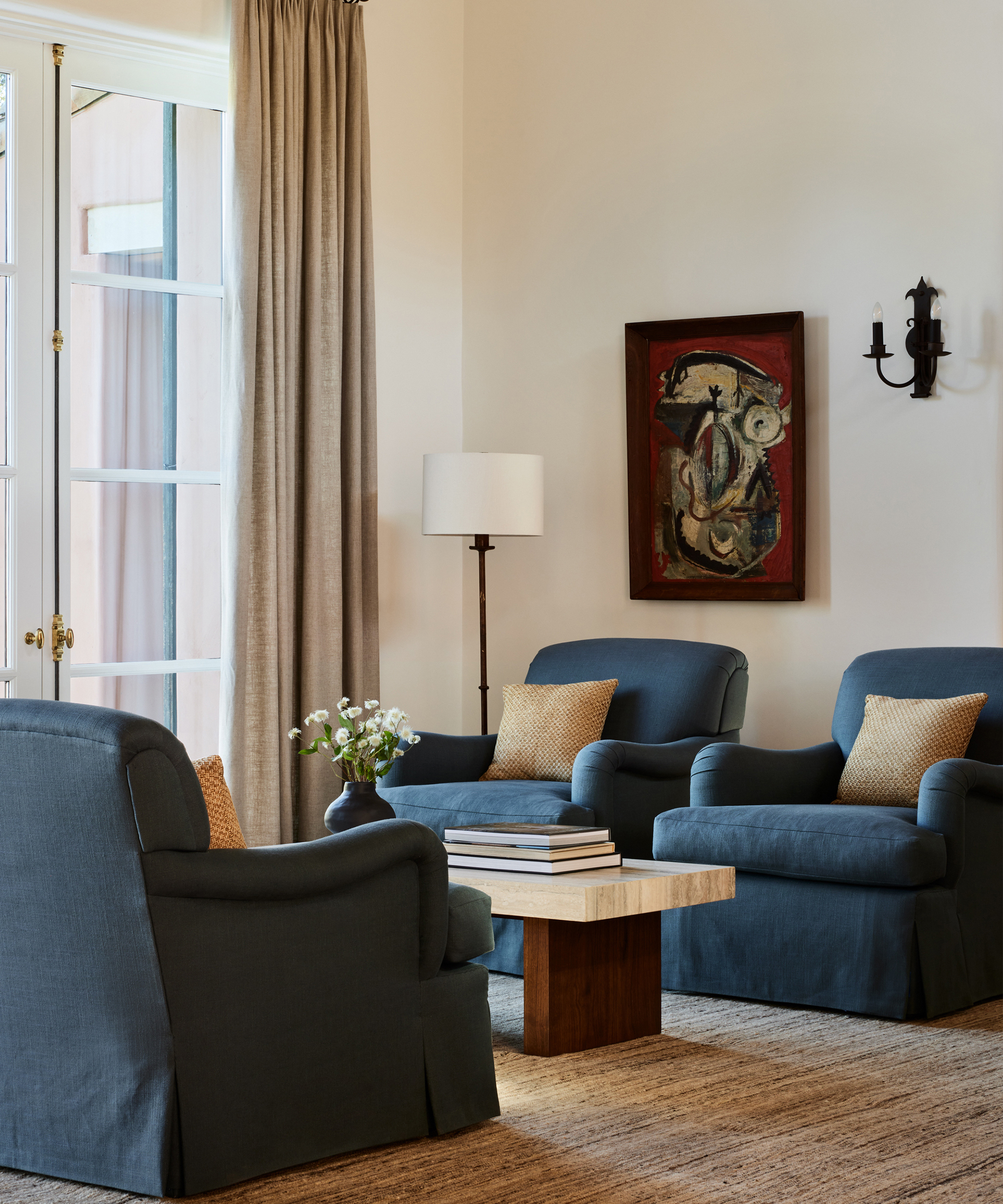 A cozy living room with two dark blue armchairs, a matching sofa, wooden coffee table with books, a floor lamp, beige curtains, abstract wall art, and neutral decor accents inspired by Corinne Mathern’s timeless style.