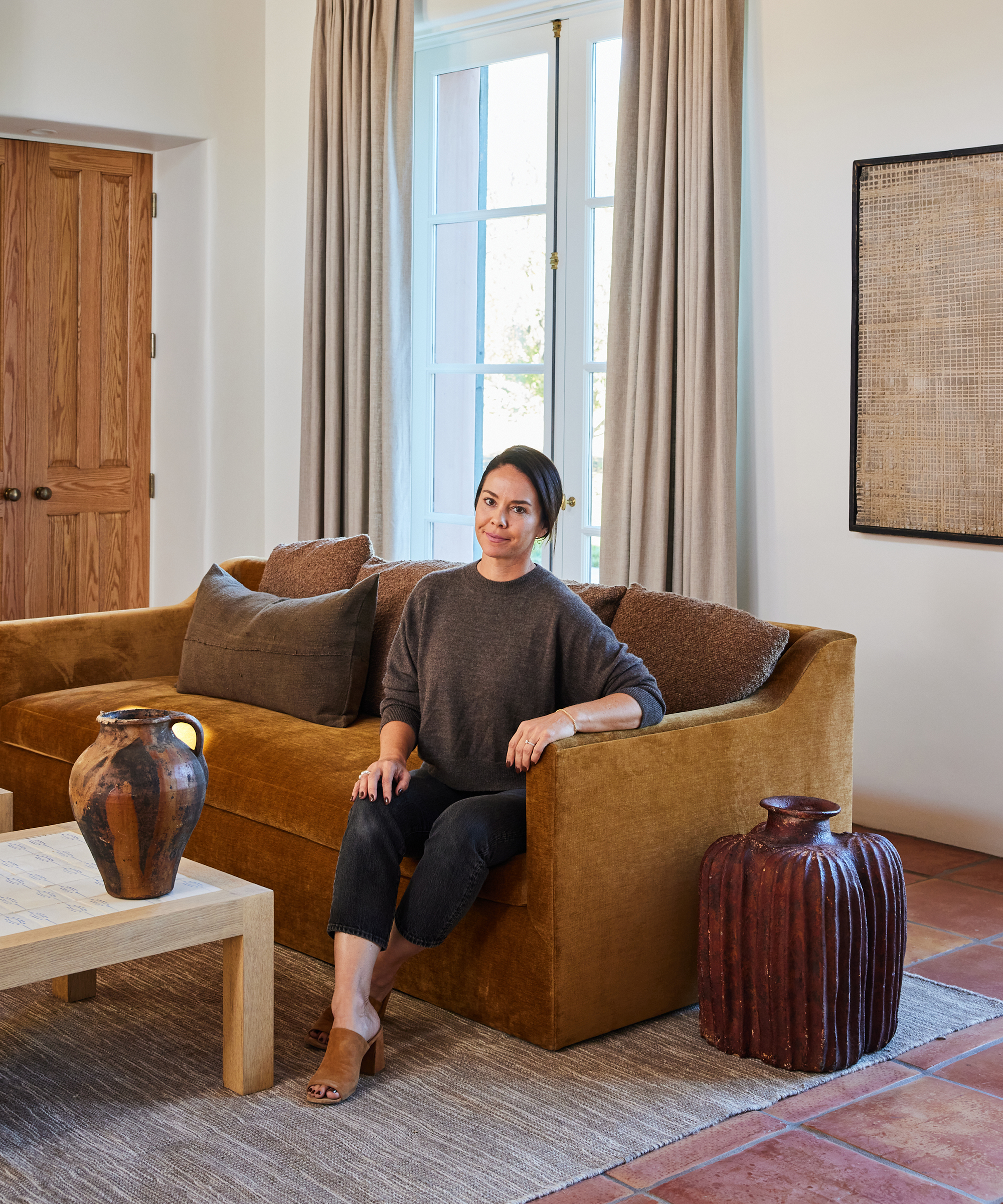 a portrait or corinne in a brown sweater and jeans sitting on a brown couch