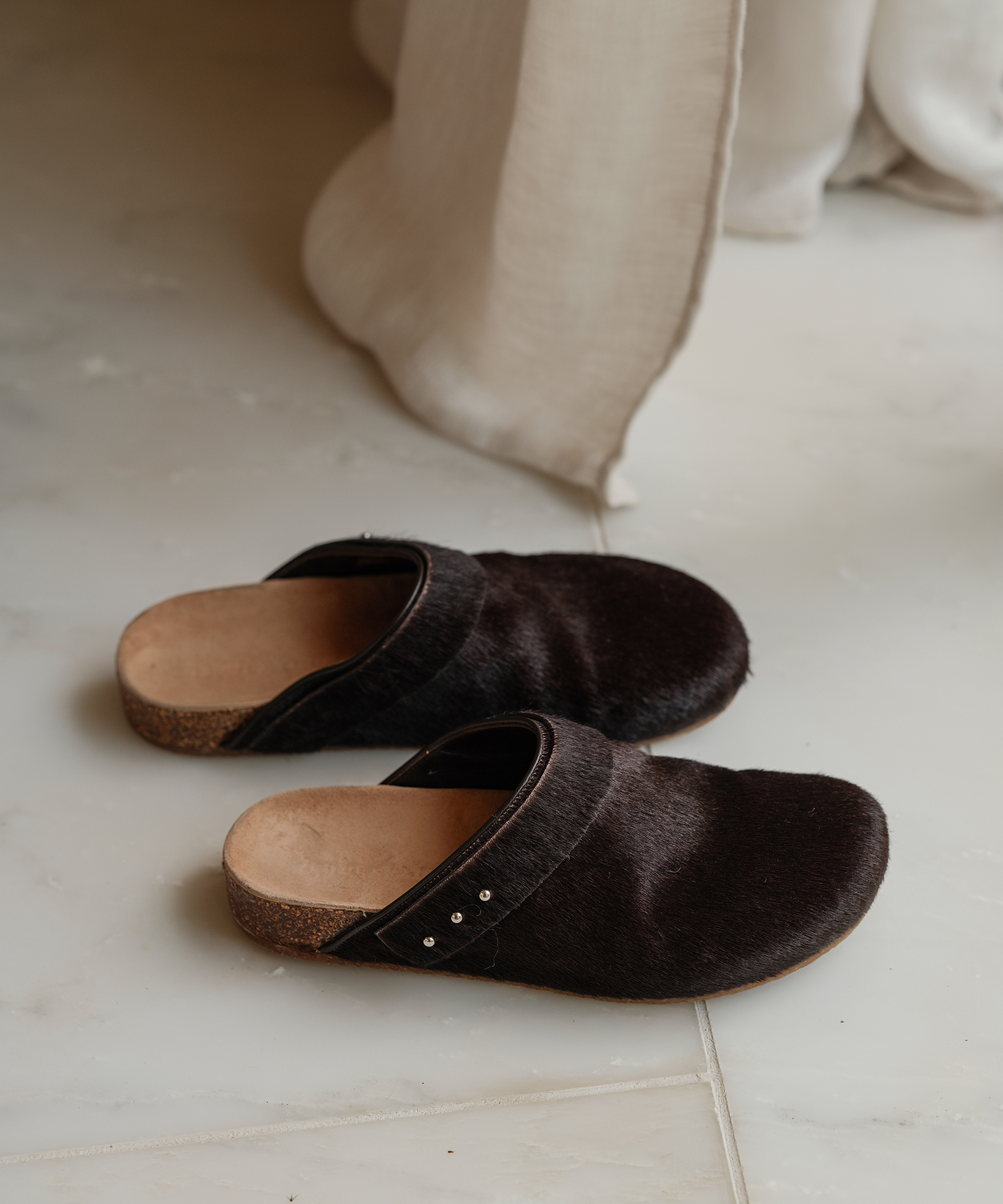 A pair of dark brown slip-on clogs with cork soles sits on a light-colored tiled floor near a white curtain, ready to pair perfectly with cozy sweaters.