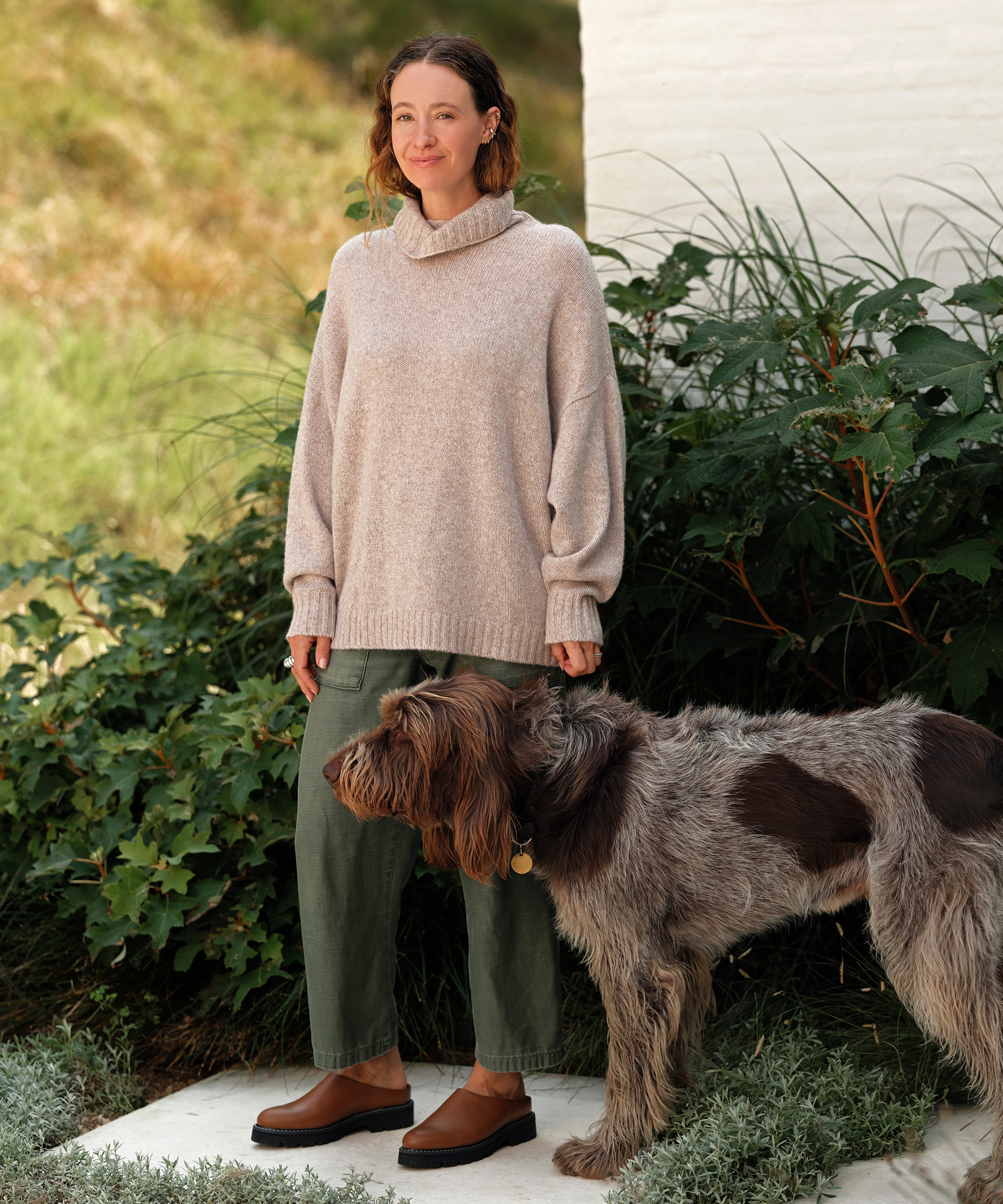 jenni standing outside on a patio in front of a grassy hill in an oatmeal cashmere jet turtleneck green pants and leather lug mules with her dog