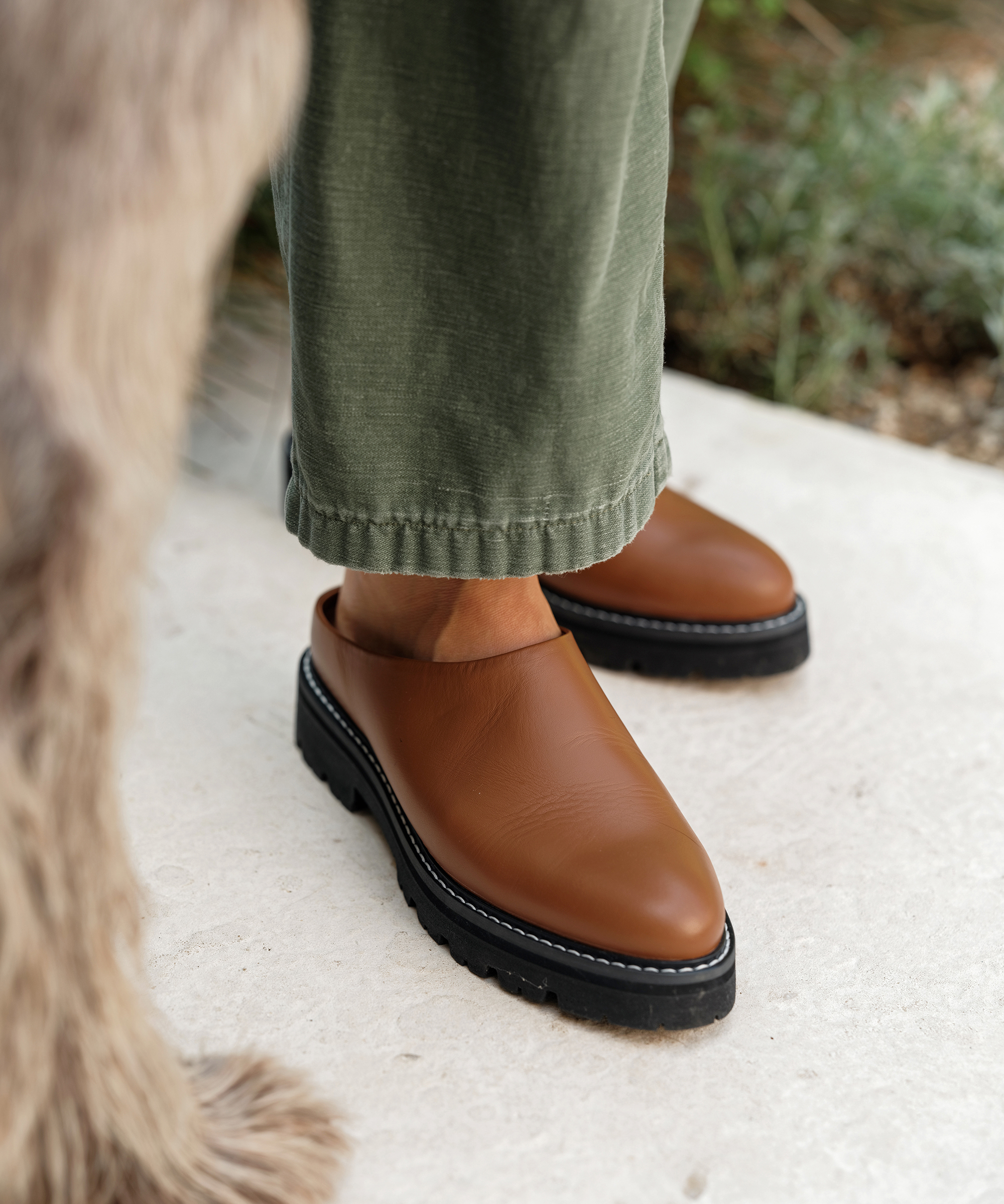A person wearing olive green pants and brown slip-on shoes with black soles stands on a light-colored surface—an inviting look for a casual Thanksgiving outfit. Part of an animal with light brown fur appears on the left side of the image.
