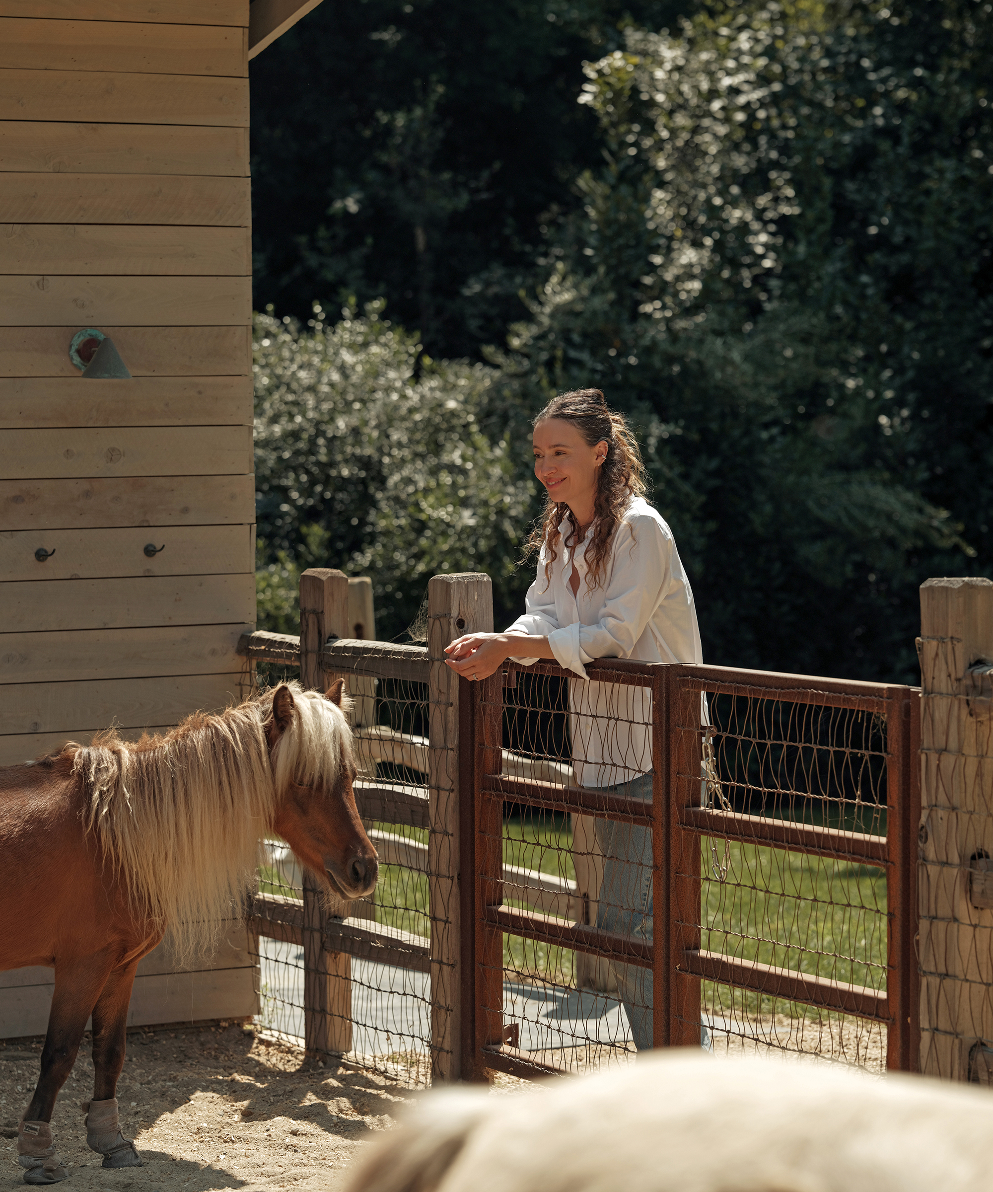 A woman with long, wavy hair stands by a wooden fence in her stylish Thanksgiving outfit, watching a small brown and white pony in a sunny outdoor enclosure beside a wooden building. Green trees fill the background.