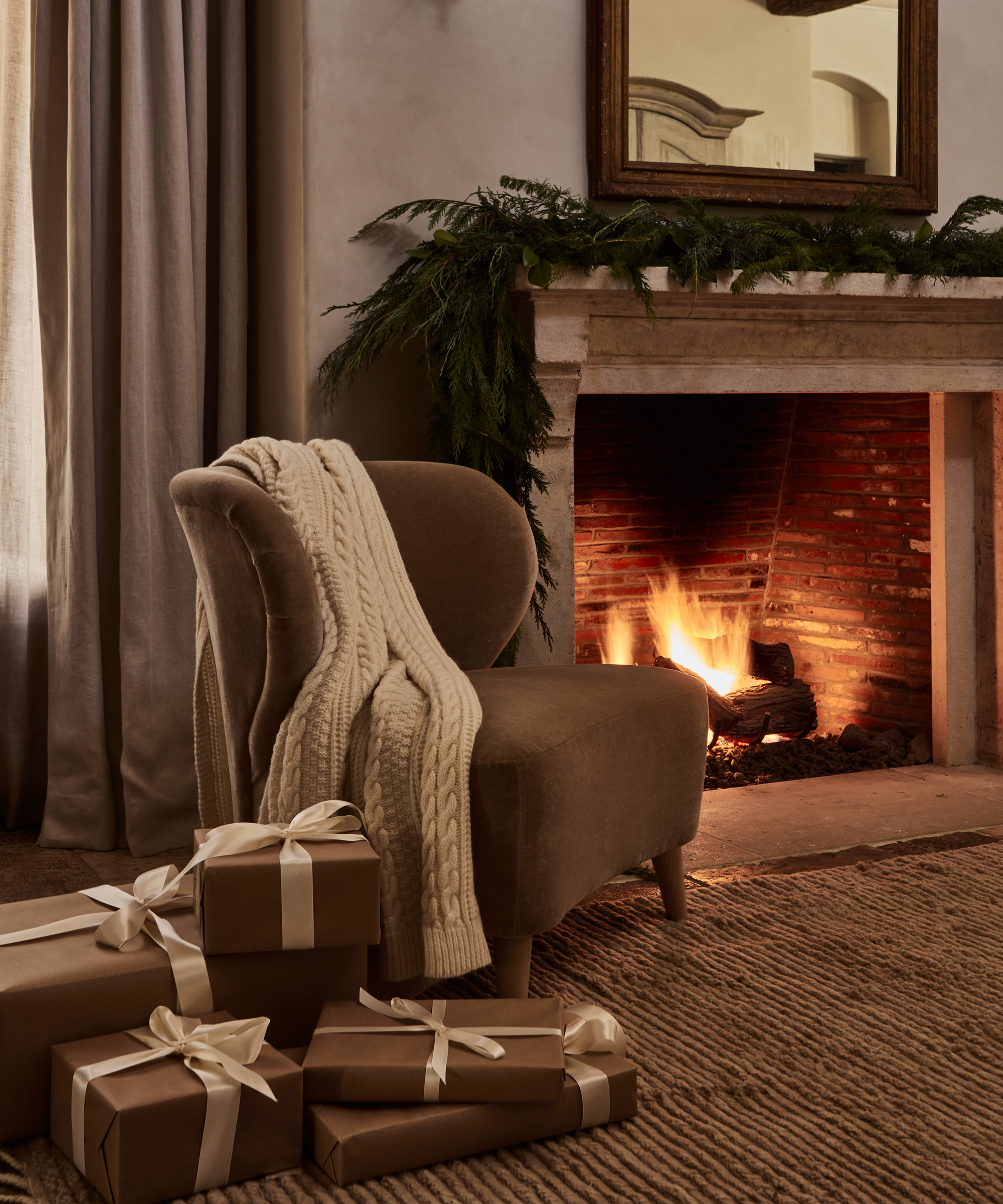 A cozy living room with a lit fireplace, a taupe chair draped with a chunky knit blanket, four brown gift boxes with white ribbons on a woven rug, and Olive Ateliers-inspired garland decor on the mantel.