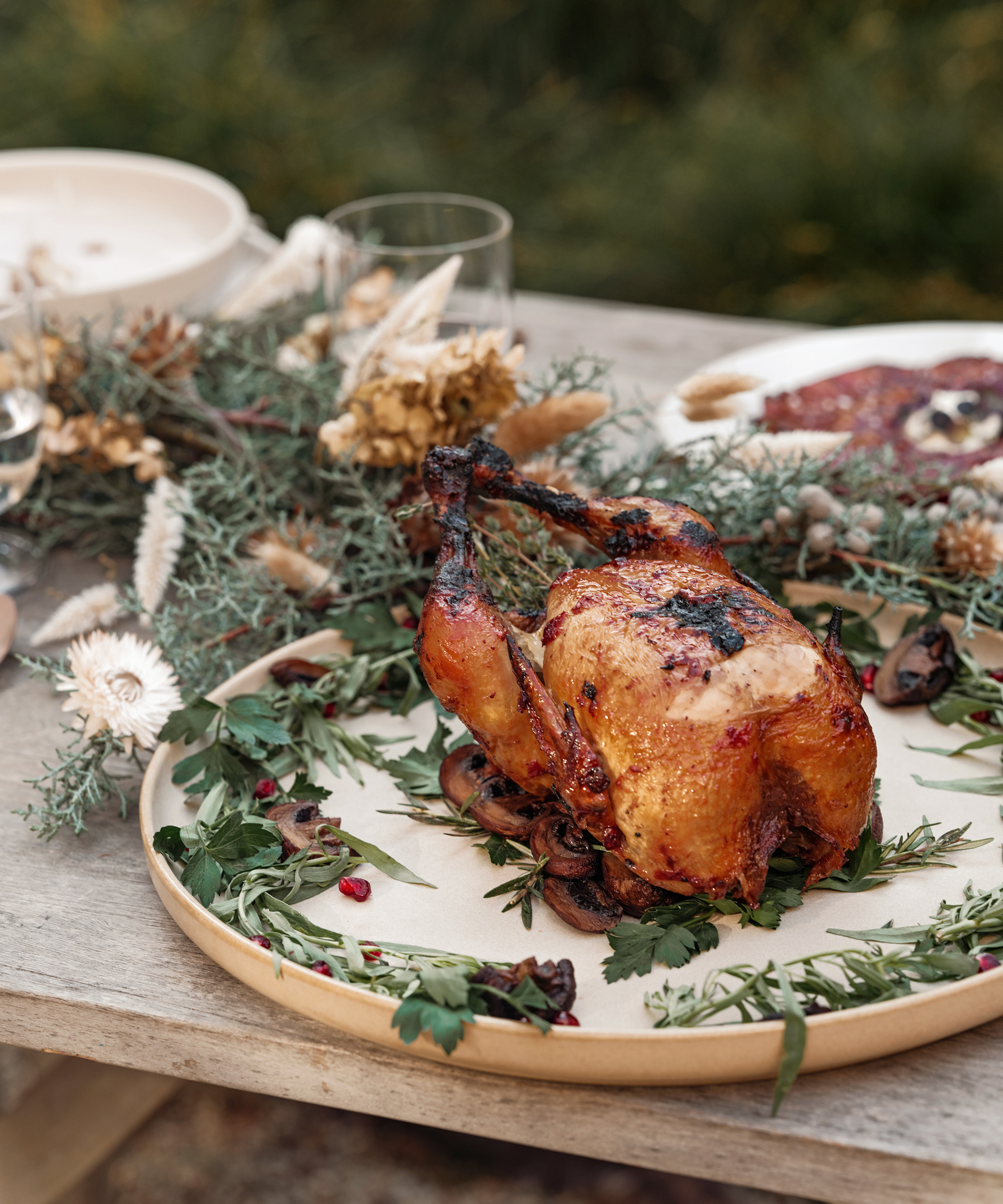 A roasted chicken garnished with herbs sits on a round plate atop a rustic wooden table, surrounded by greenery, dried flowers, and elegant tableware set for a festive holiday dinner.