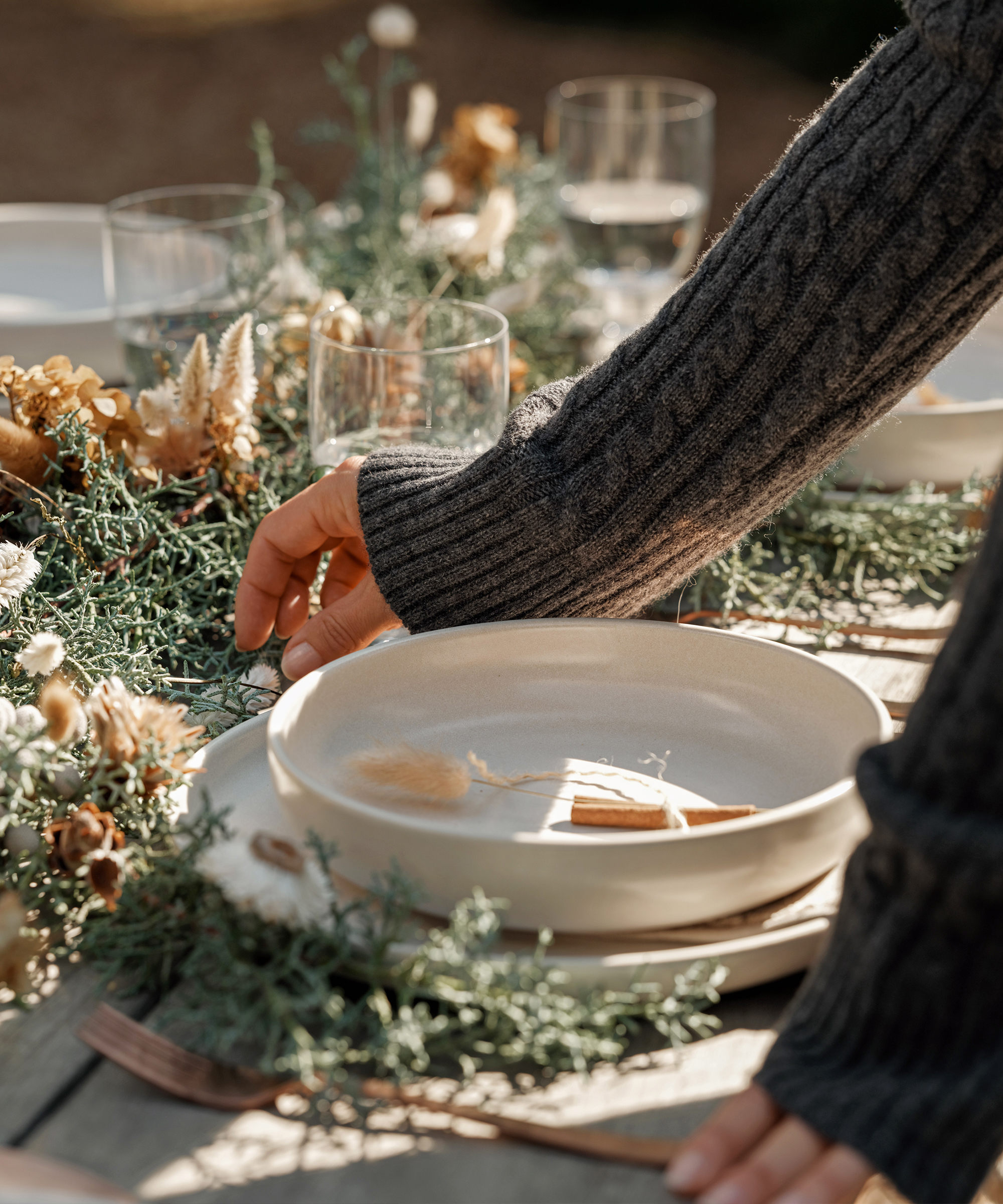 A person in a gray cable-knit sweater adjusts a beige ceramic bowl on an outdoor table set for a holiday dinner, decorated with dried flowers, greenery, and glassware in natural sunlight.