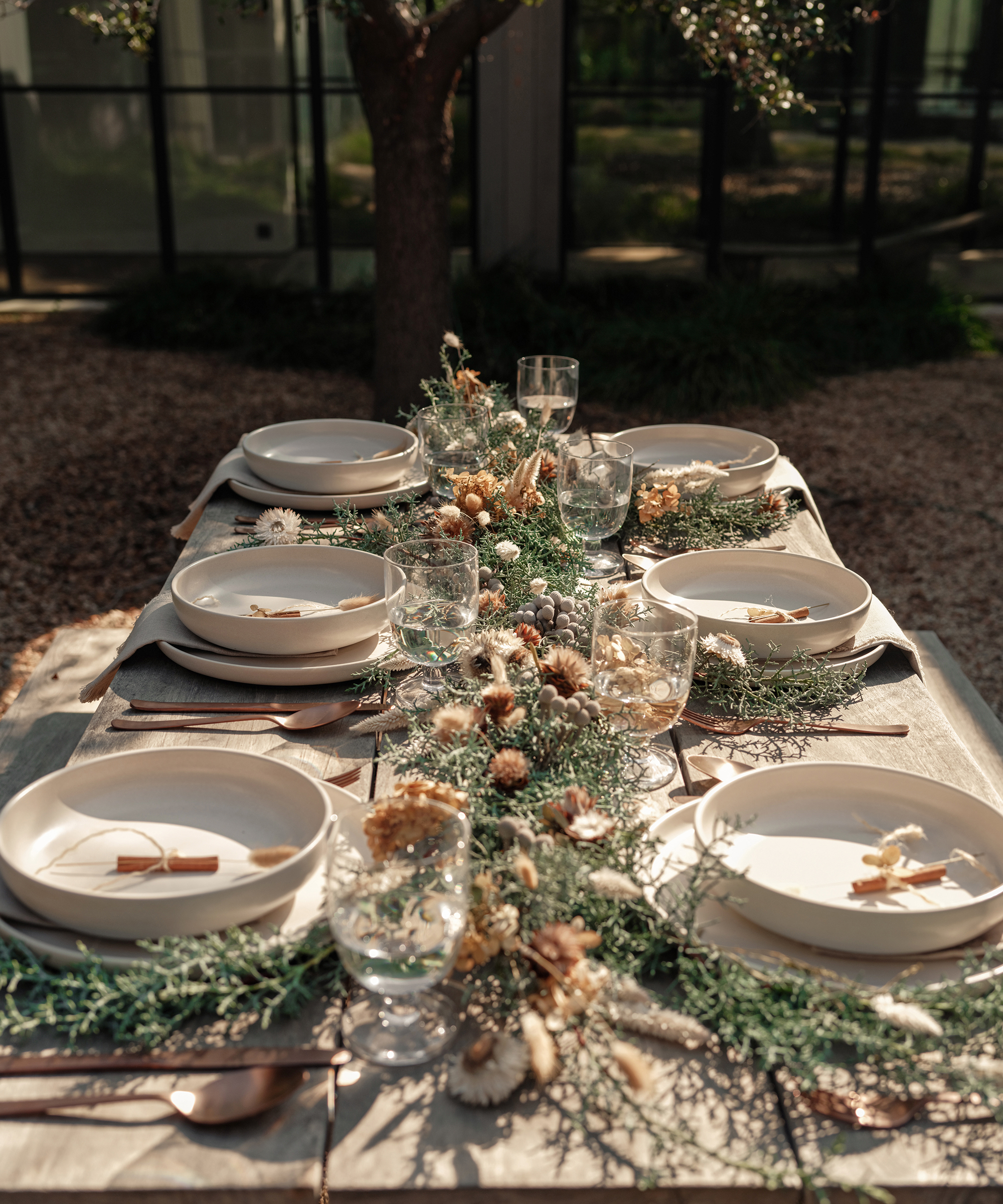 A sunlit outdoor dining table set for six with white plates, glassware, and silverware evokes a welcoming holiday dinner. The centerpiece of greenery and dried flowers adds a natural, rustic touch as soft sunlight highlights the elegant arrangement.