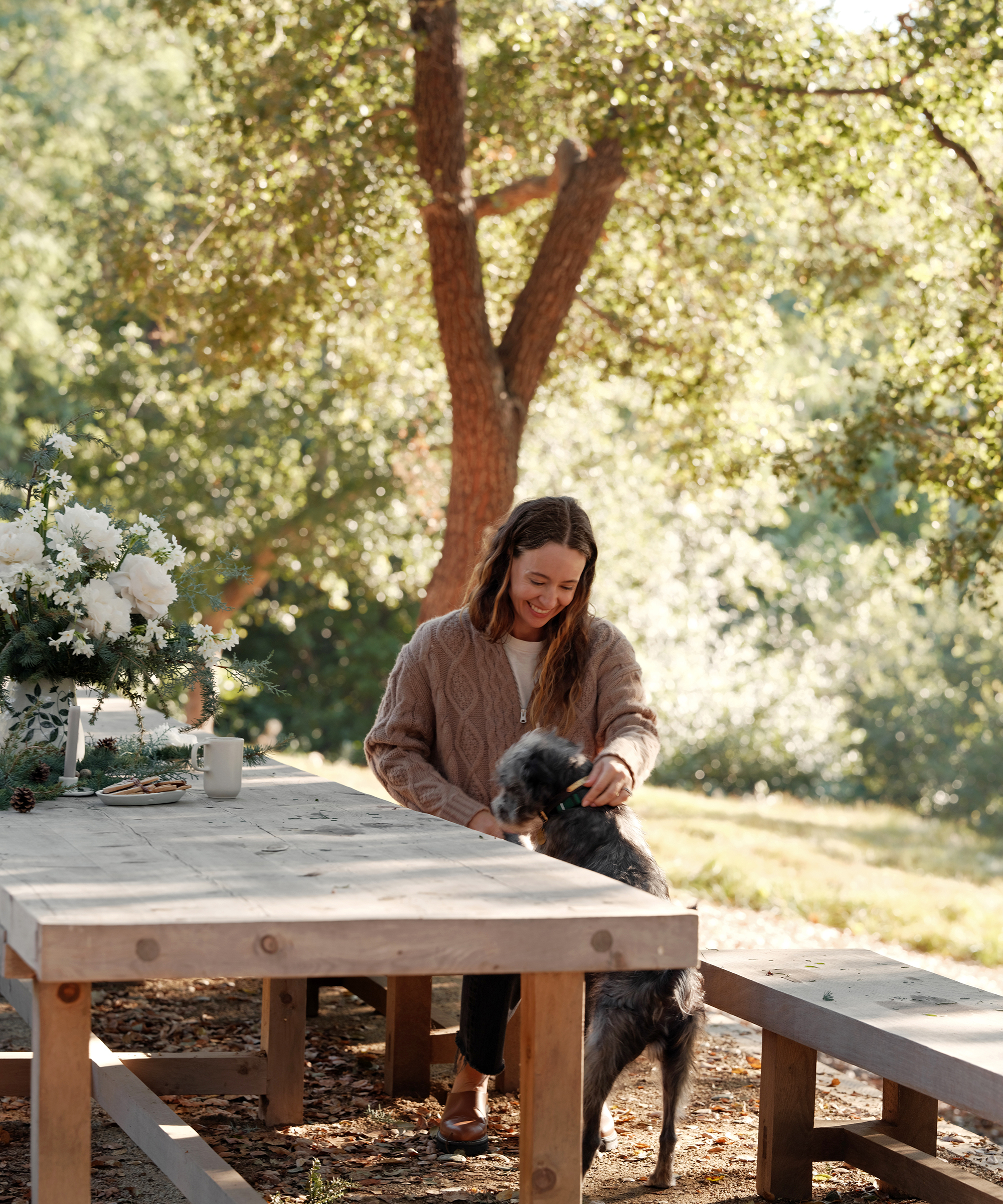 A woman in a cozy sweater sits at a wooden picnic table outdoors, smiling and petting a gray dog beside her. Sunlight filters through the trees, and easy holiday desserts sit next to a vase of white flowers on the table.