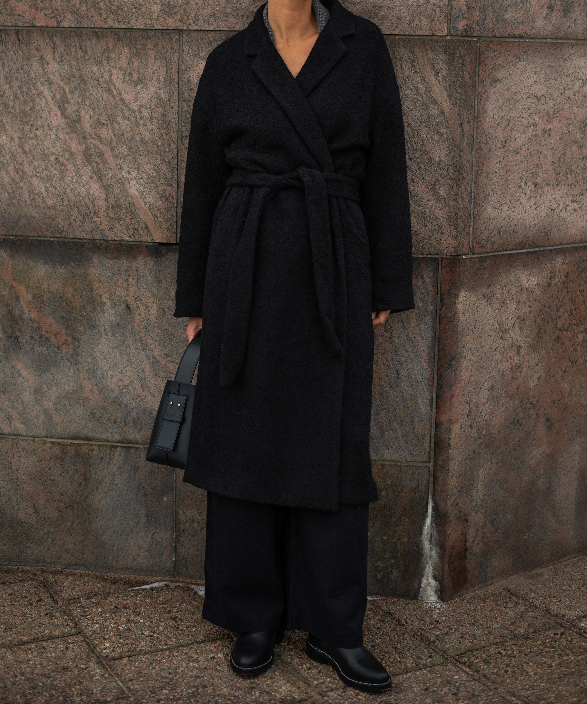 a woman wearing a black coat and black leather bag leaning against a stone wall