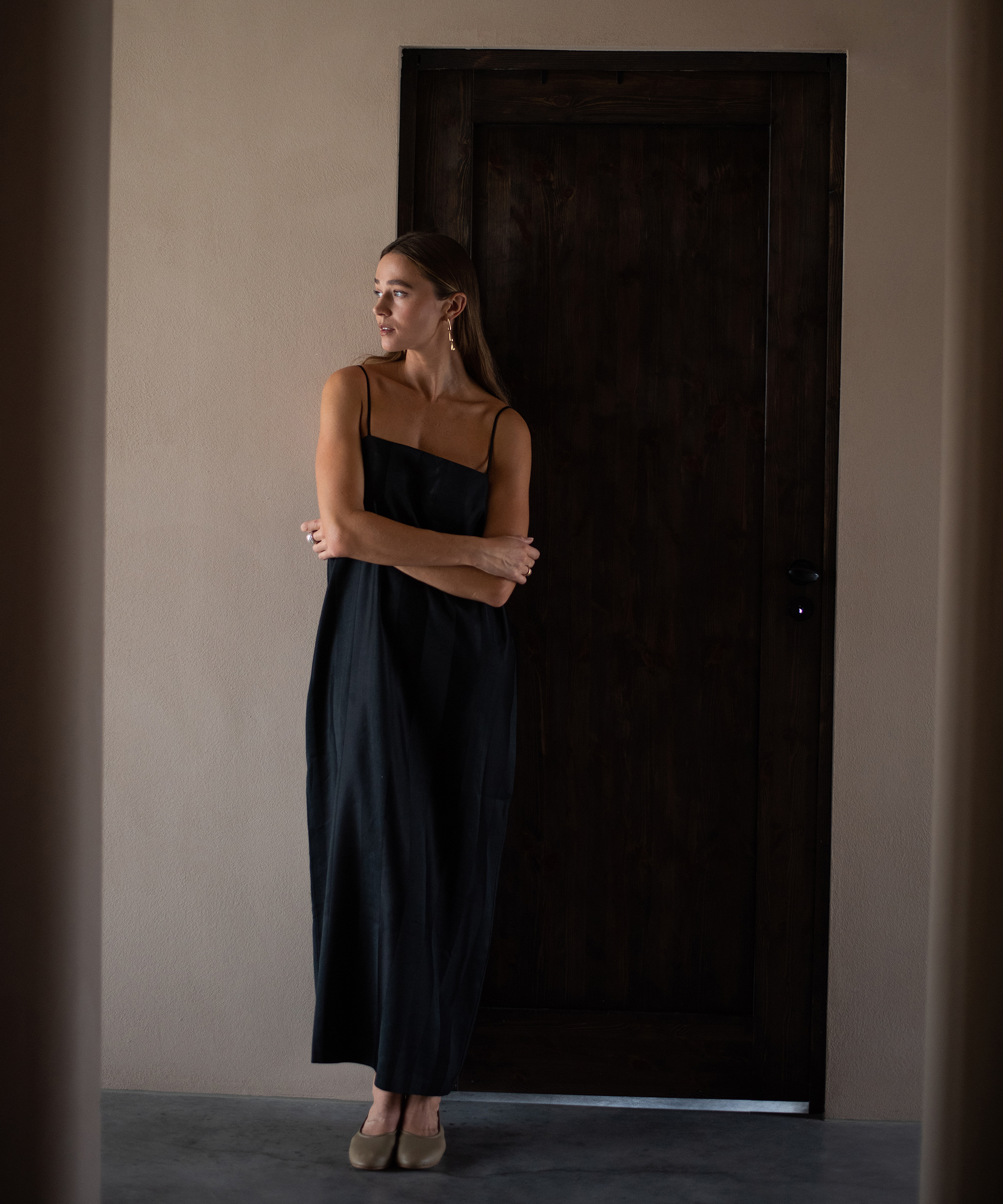A woman in a black sleeveless dress stands barefoot with her arms crossed, leaning against a dark wooden door in a softly lit room, looking to her left.